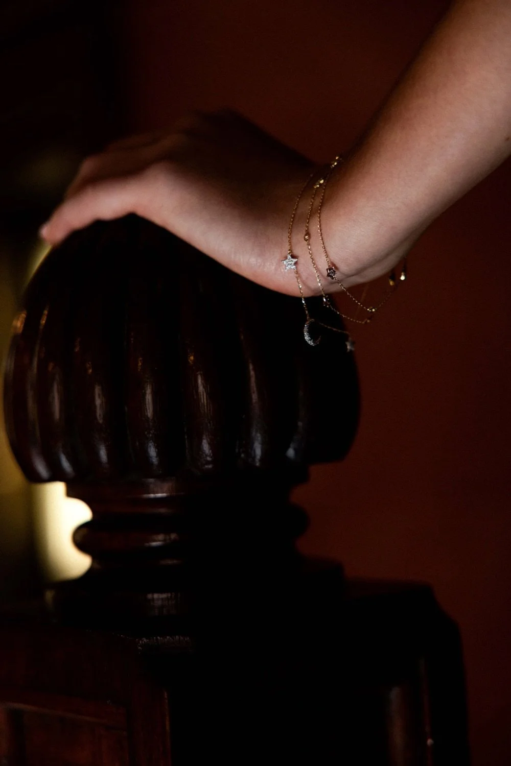A person's hand resting on a dark wooden table, adorned with delicate gold and silver star and moon necklaces.