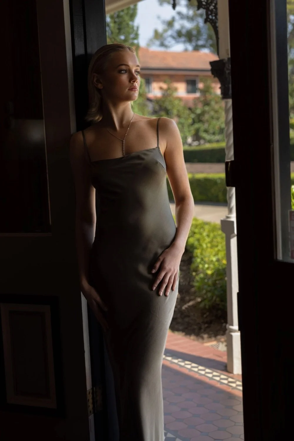 A woman standing in a doorway, looking outdoors. She is wearing a silky, spaghetti strap dress and a necklace. The scene outside is green with trees and a building in the background.