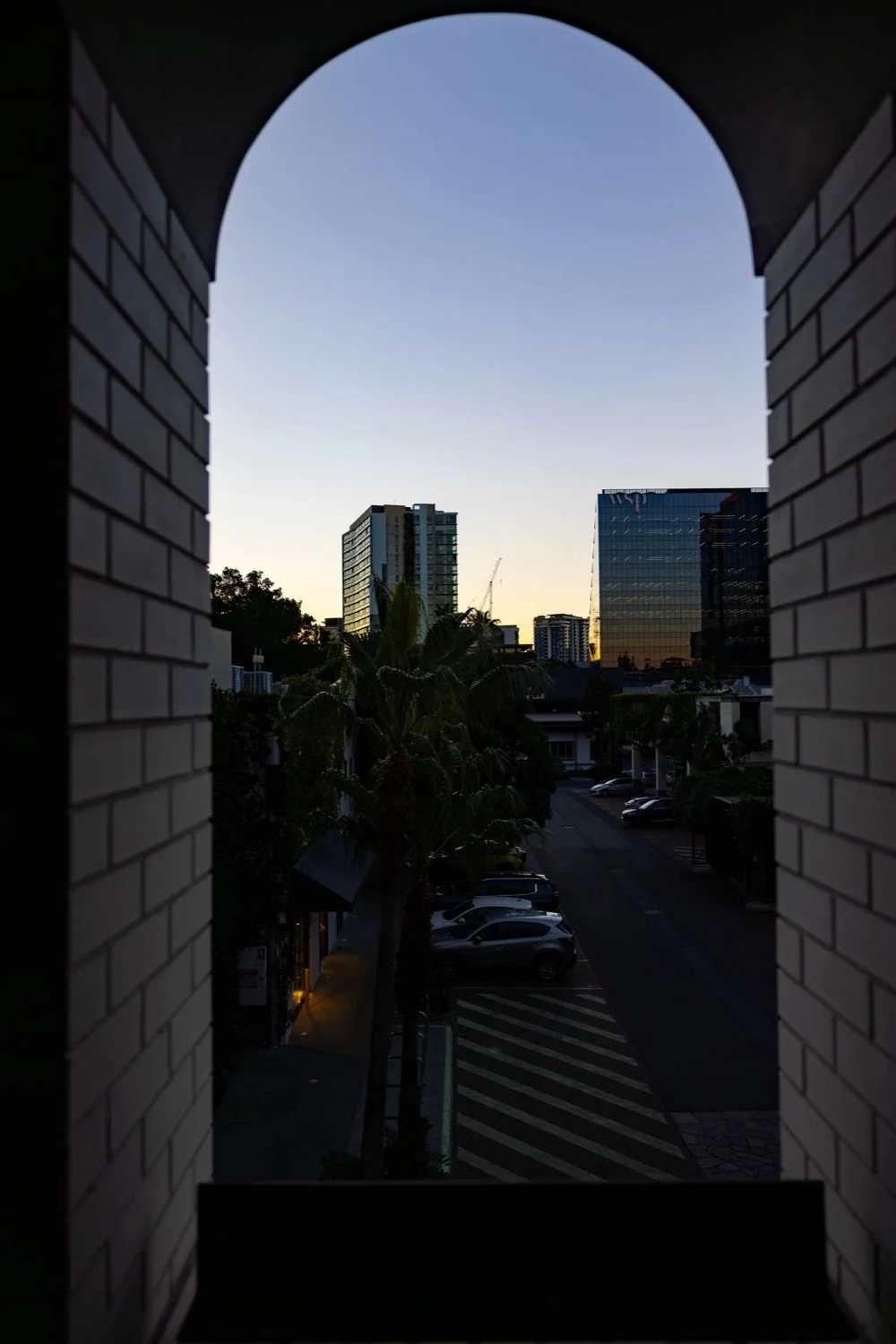 View of a cityscape seen through an arched window, featuring tall modern buildings, a construction crane, palm trees, and a parking lot at dusk.