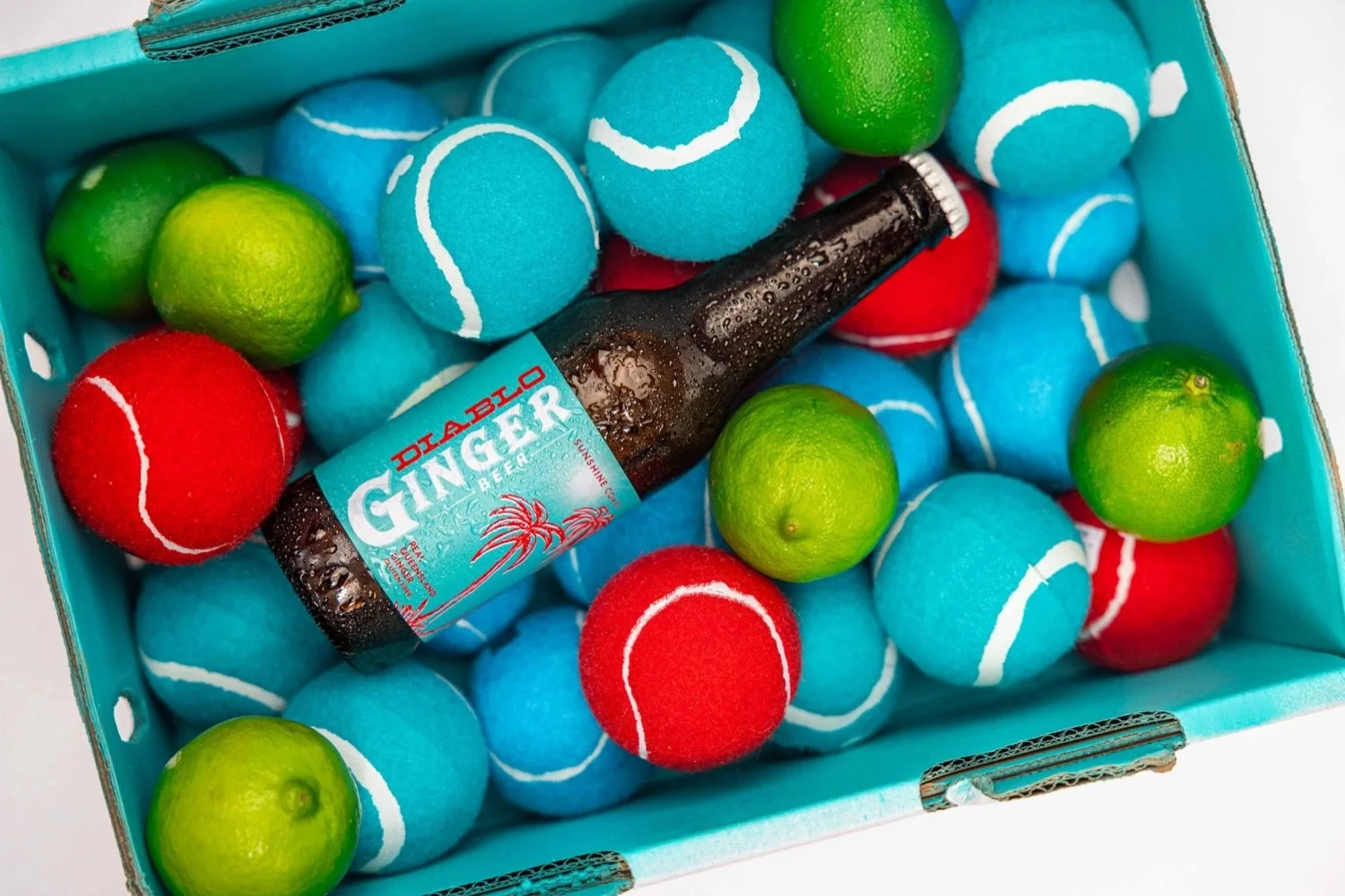 A box of tennis balls in red, blue, and green, with a small bottle of Delabelo Ginger Beer surrounded by green limes inside the box.