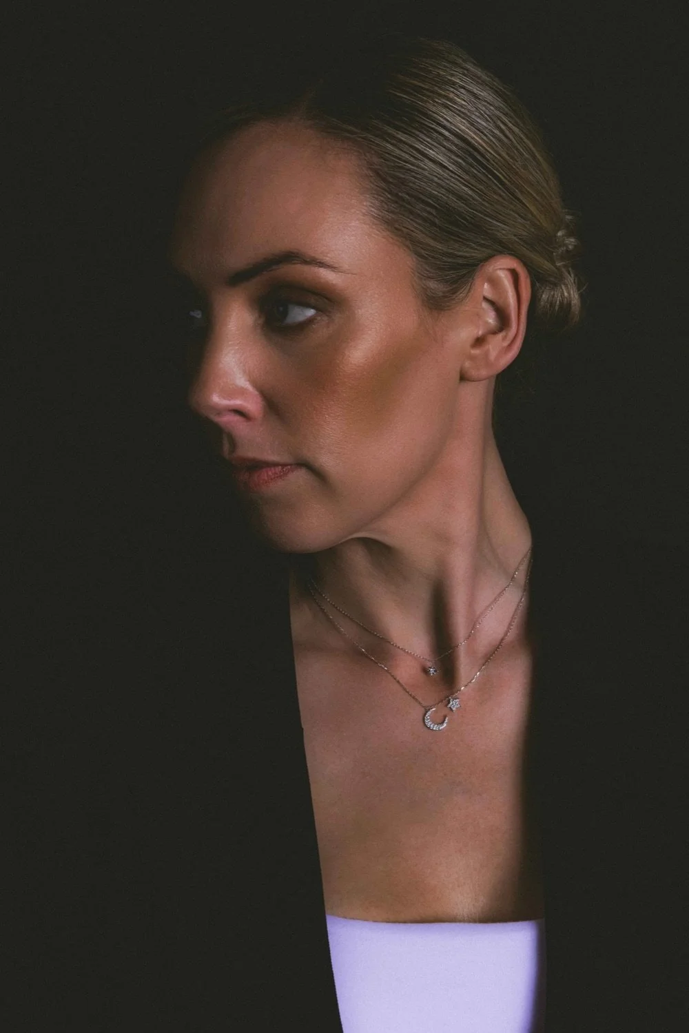 Close-up of a woman with blonde hair styled in a bun, wearing multiple layered necklaces, including a crescent moon pendant, against a dark background.