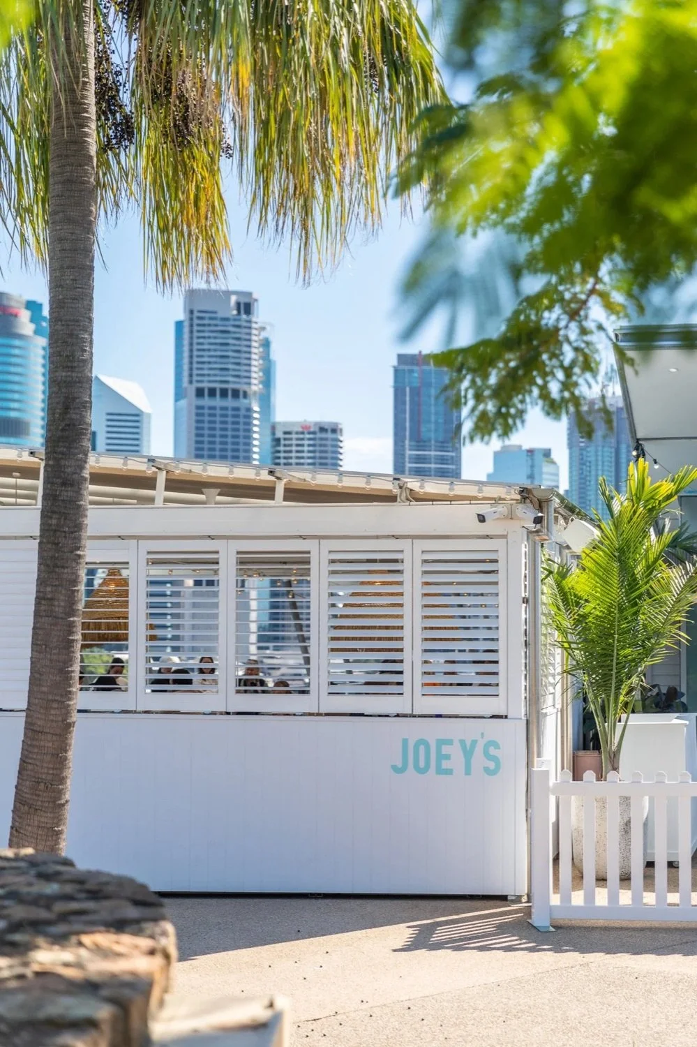 Outdoor view of a white structure with the sign 'Joey's' in blue, surrounded by palm trees, with city skyscrapers in the background, under a clear blue sky.
