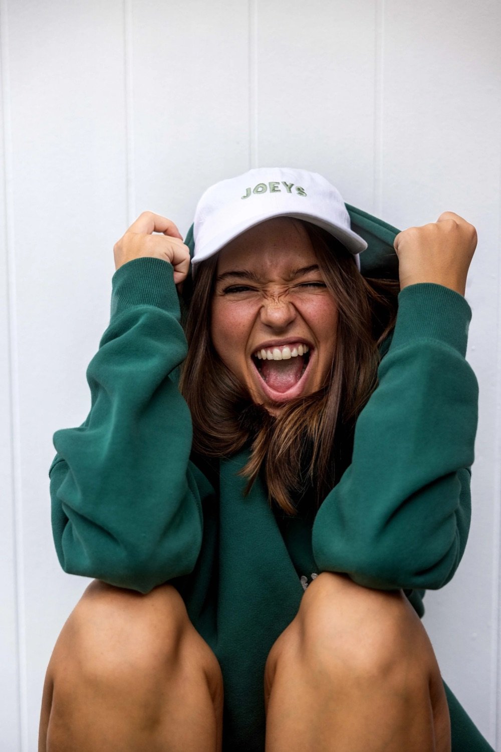 A woman with long brown hair wearing a green sweatshirt and white cap that says 'JOEYS' is sitting with her knees up, smiling with her mouth open and fists raised near her head, expressing excitement or joy.