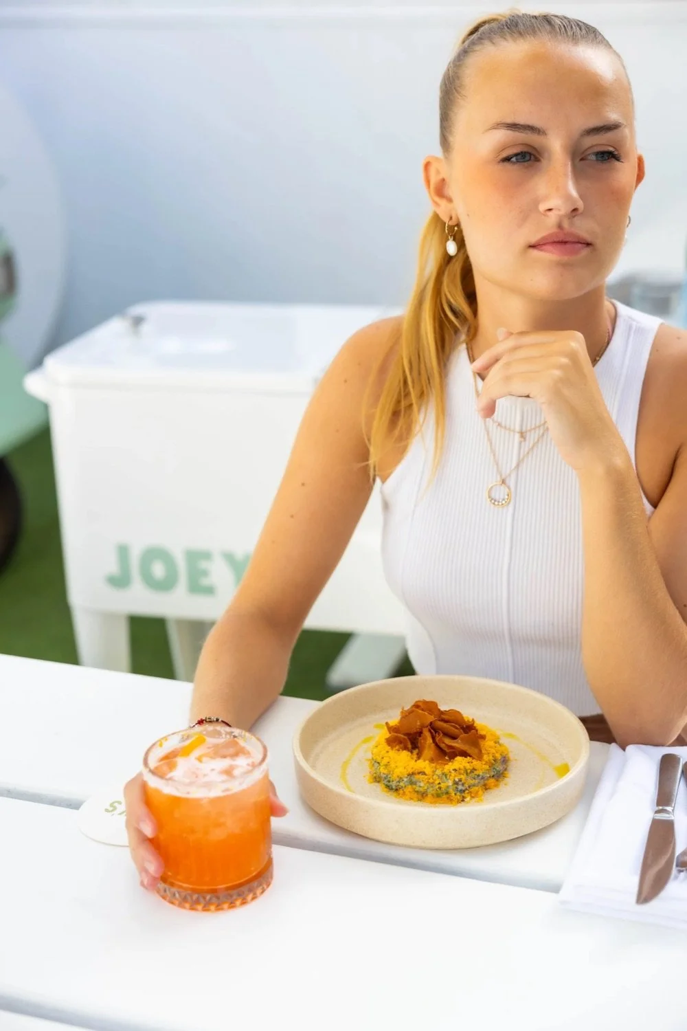 A woman with blonde hair tied back, wearing a white sleeveless top and jewelry, sits at a table with a beige bowl of food and a glass of orange-colored drink. She is resting her chin on her hand and looking contemplative. Background includes a white 