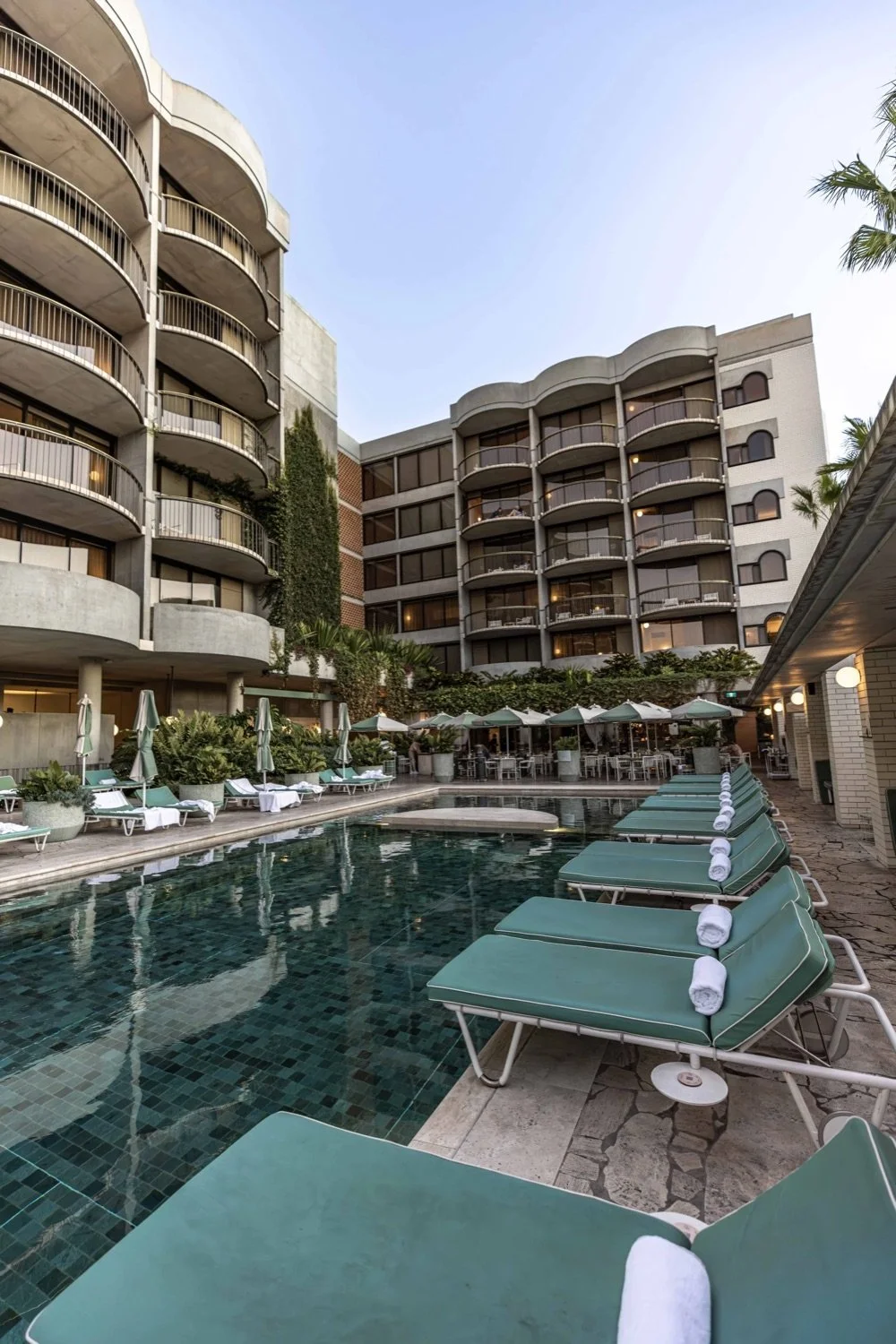 Swimming pool area with lounge chairs and umbrellas in a hotel courtyard, surrounded by multi-story hotel buildings with balconies.