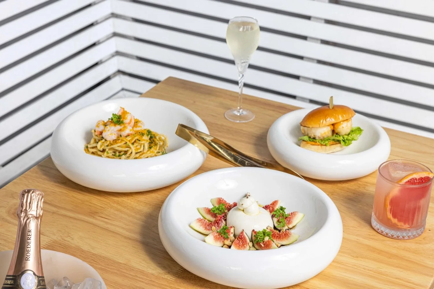 A table with various dishes including a shrimp pasta, a sandwich with shrimp, a dessert with figs and a cream topping, a glass of champagne, a pink cocktail, and a bottle of rosé champagne, set against a black and white striped background.