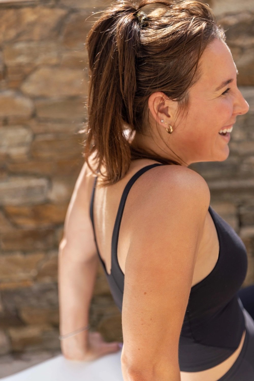 A smiling woman with shoulder-length brown hair wearing a black tank top and gold hoop earrings, sitting against a rustic wooden wall.