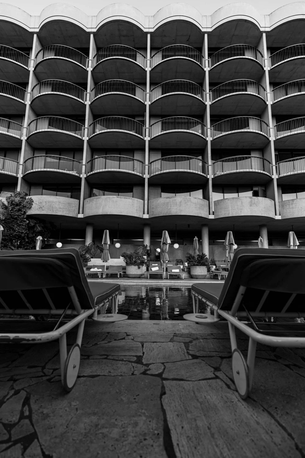 A black-and-white photograph of a hotel or apartment with multiple balconies, poolside lounge chairs, umbrellas, and potted plants.