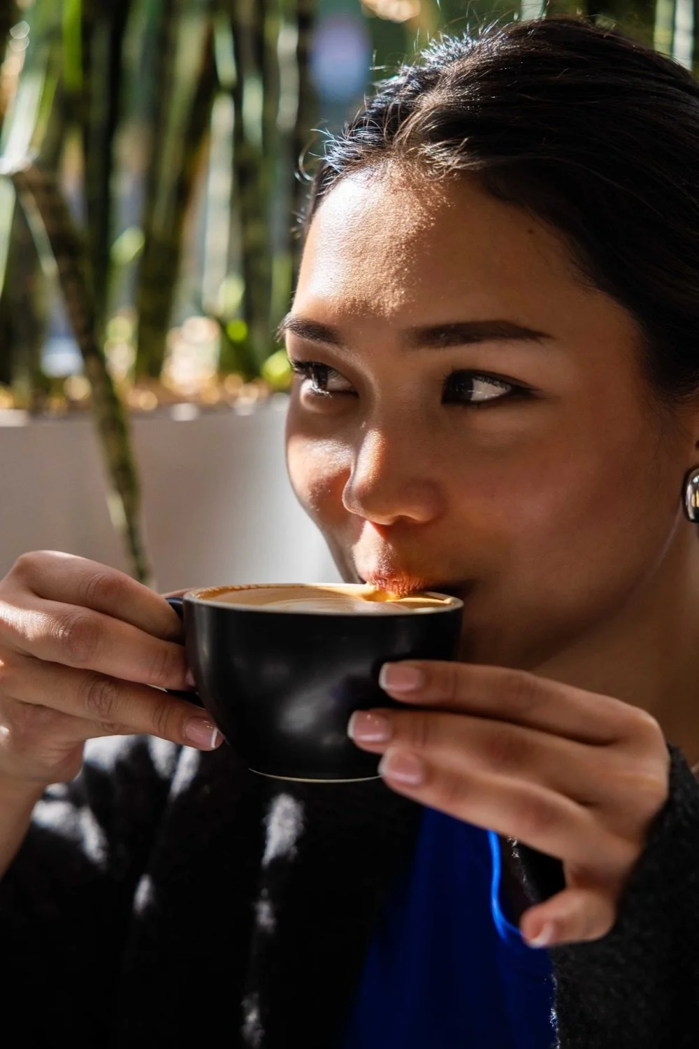 A woman with dark hair, wearing earrings, is holding a black cup of coffee and taking a sip, with sunlight highlighting her face and a blurred background of green plants.