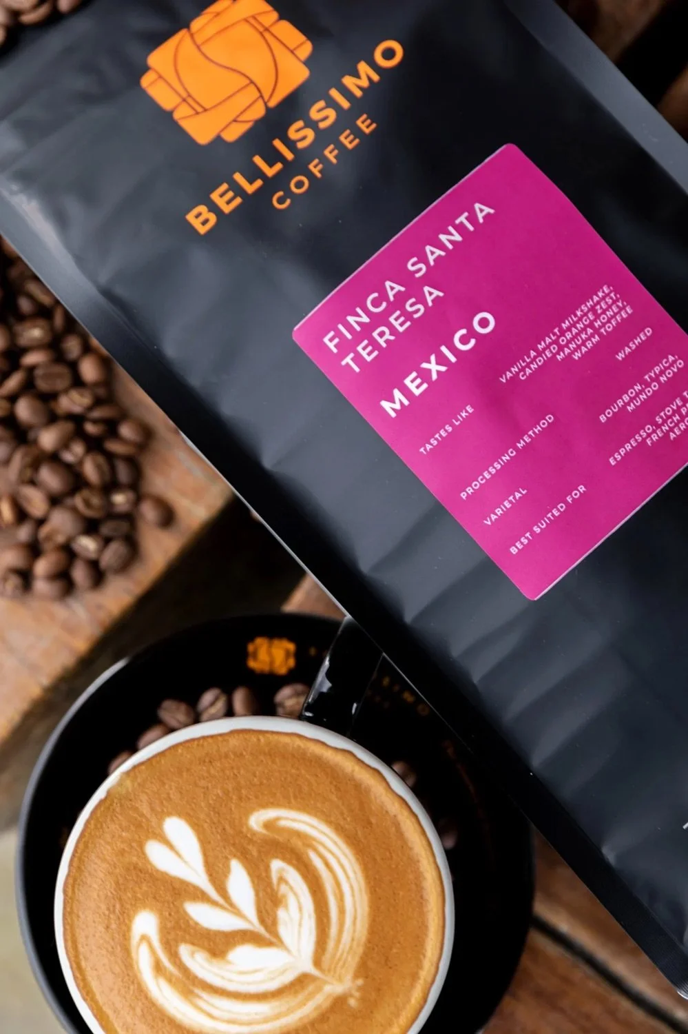 A black bag of Bellissimo Coffee with Finca Santa Teresa Mexico label next to a cup of latte with latte art, surrounded by coffee beans on a wooden surface.