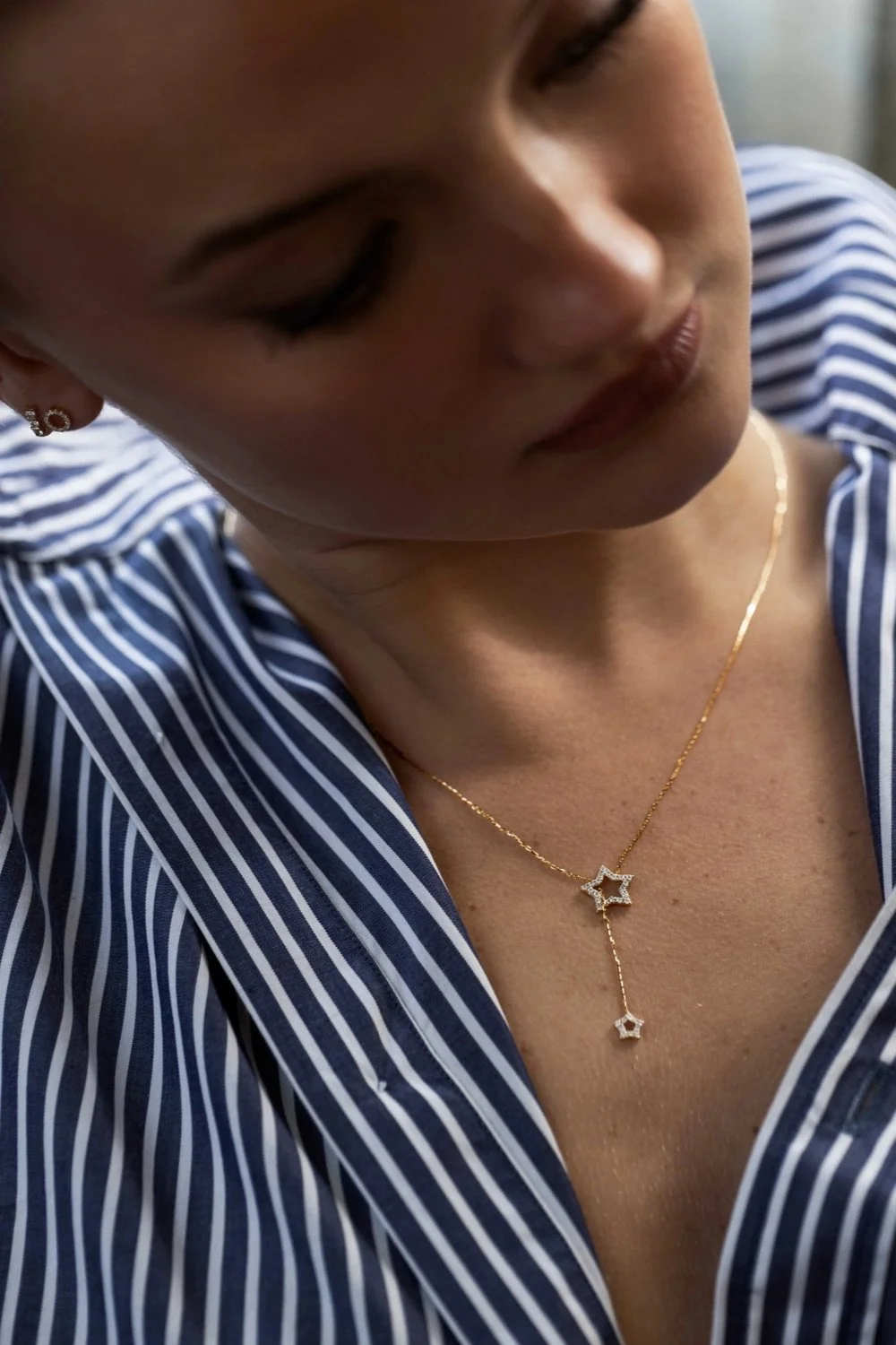 Close-up of woman wearing a striped shirt and a delicate necklace with star-shaped charms, her face turned downward with eyes closed.