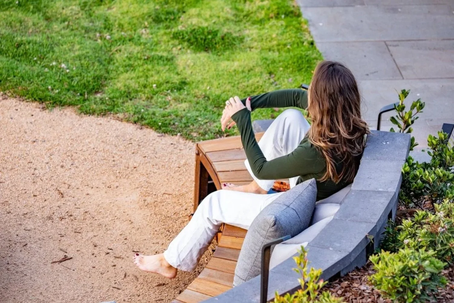 A woman sitting on a wooden and stone outdoor bench, with her feet on the ground and her arms resting on her knees, gazing into the distance in a park setting.