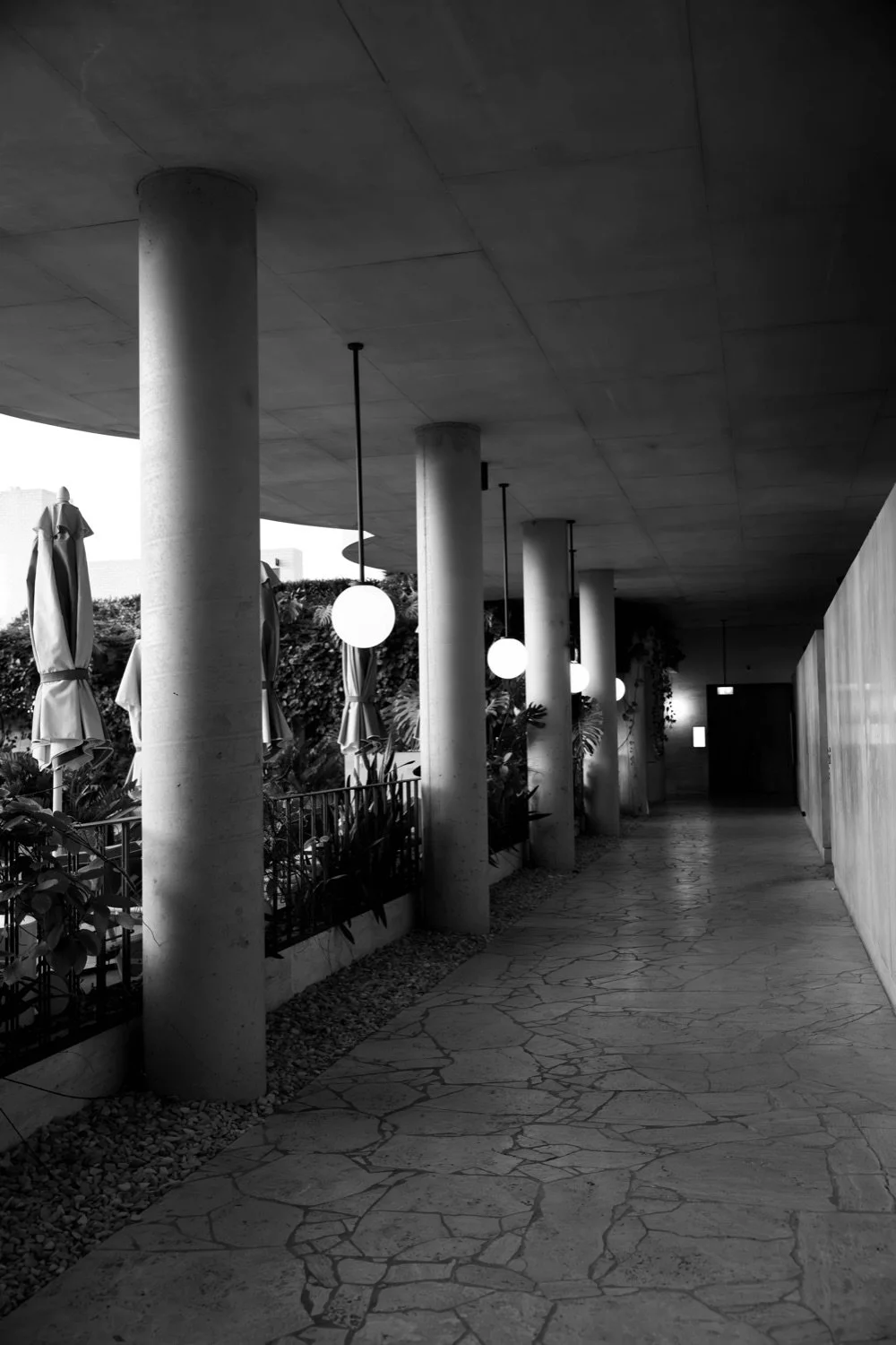 A black and white photo of an outdoor corridor with large concrete pillars, round hanging lights, potted plants, and closed umbrellas. The corridor has a textured stone floor and an overhanging ceiling.