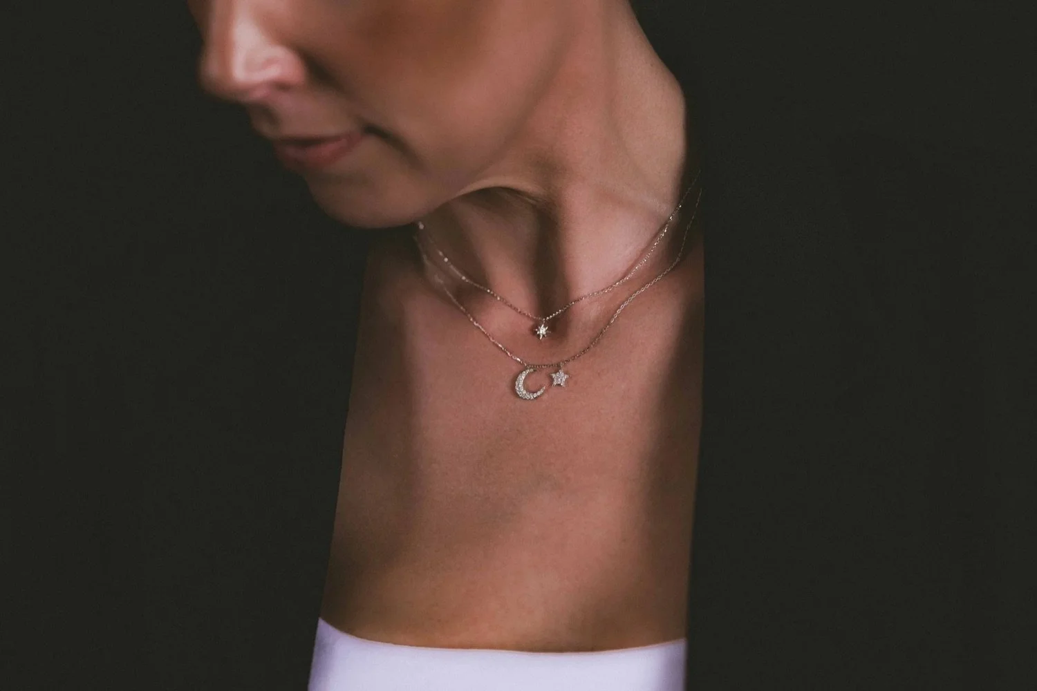 Close-up of a woman wearing layered necklaces with star, crescent moon, and starball pendants, dressed in a black top and white bottom.