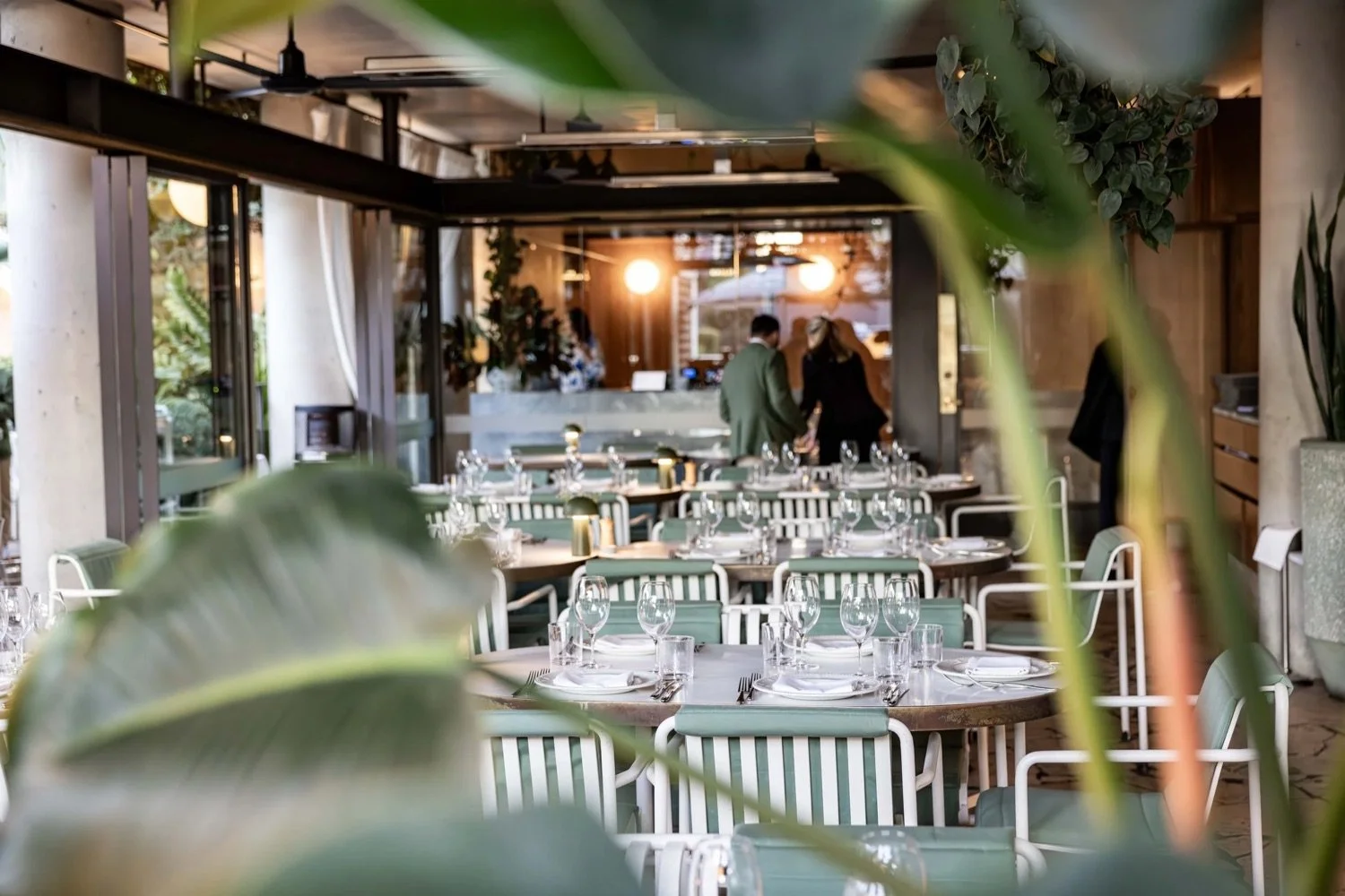 Inside a restaurant with table settings, glassware, and cutlery, viewed through large leaves, with two people standing at the back near the bar area.