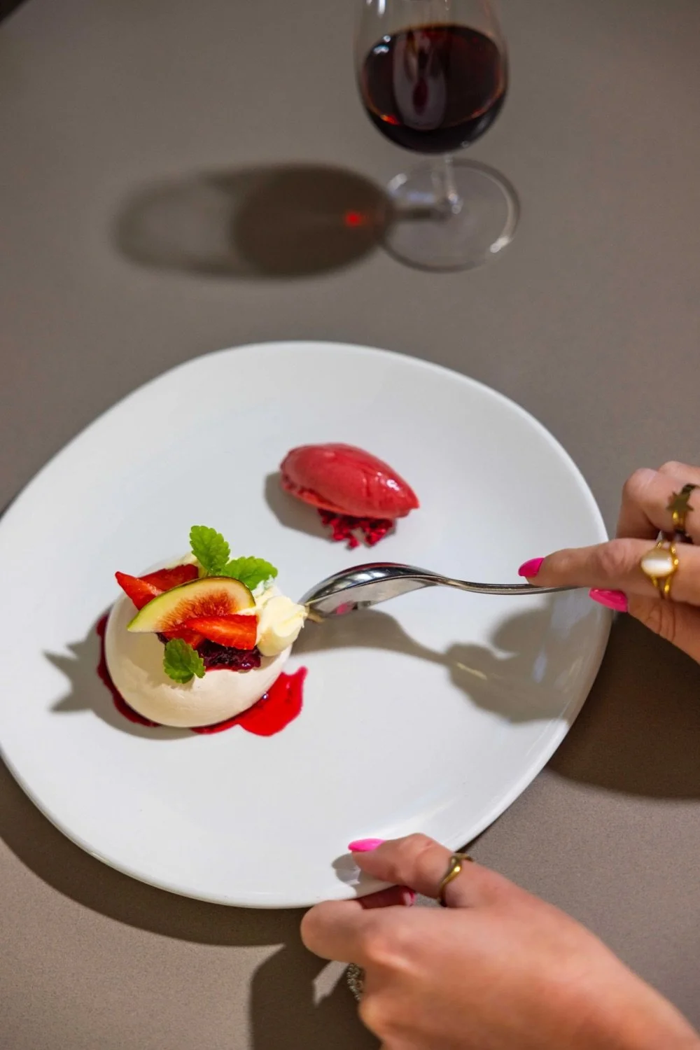 A white plate with a dessert topped with strawberries, a slice of fig, and mint leaves, alongside a scoop of pink ice cream and a glass of red wine in the background.