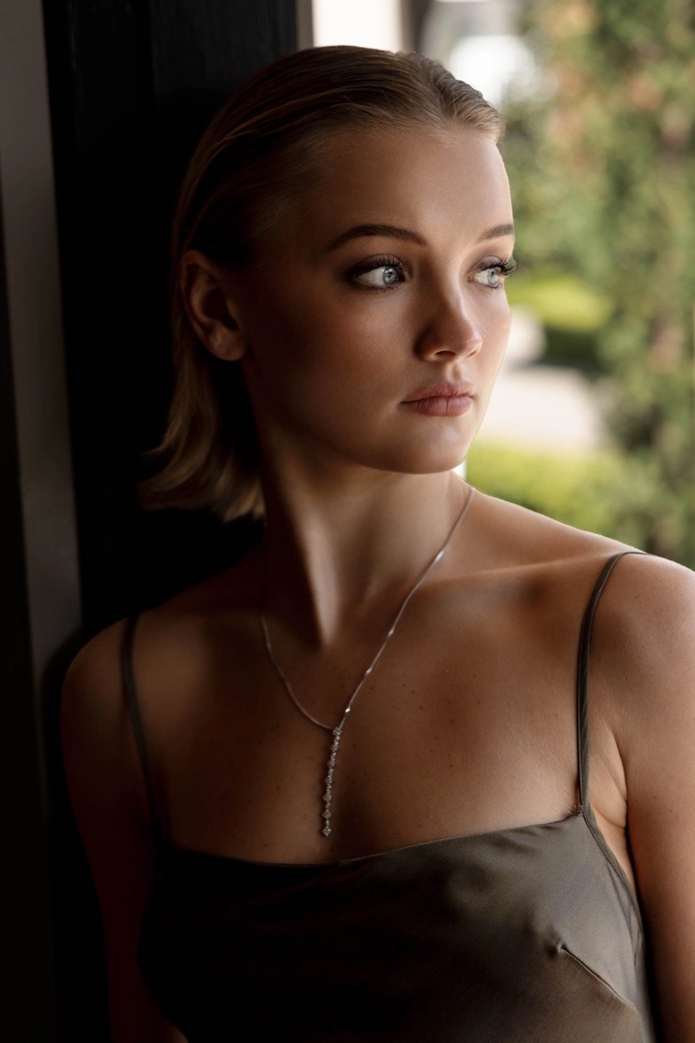 A young woman with blonde hair styled in a bob, wearing a satin spaghetti strap top and a delicate necklace, standing near a window and looking outside with a thoughtful expression.