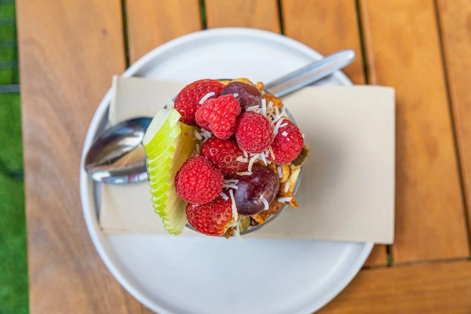 A cup of frozen yogurt topped with strawberries, raspberries, cherries, sliced apple, shredded coconut, and other toppings, on a white plate with a spoon and napkin on a wooden table.