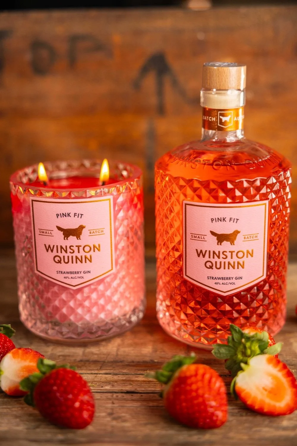 A pink textured candle with two wicks lit, placed beside a textured bottle of Winston Quinn Strawberry Gin, surrounded by fresh strawberries on a wooden surface.