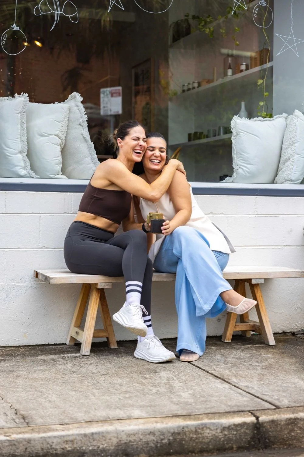 Two women sitting on a wooden bench outside a café, laughing and hugging each other. One woman is wearing a black sports bra and black leggings, and the other is in a white top and blue pants. They appear to be enjoying a happy moment together.