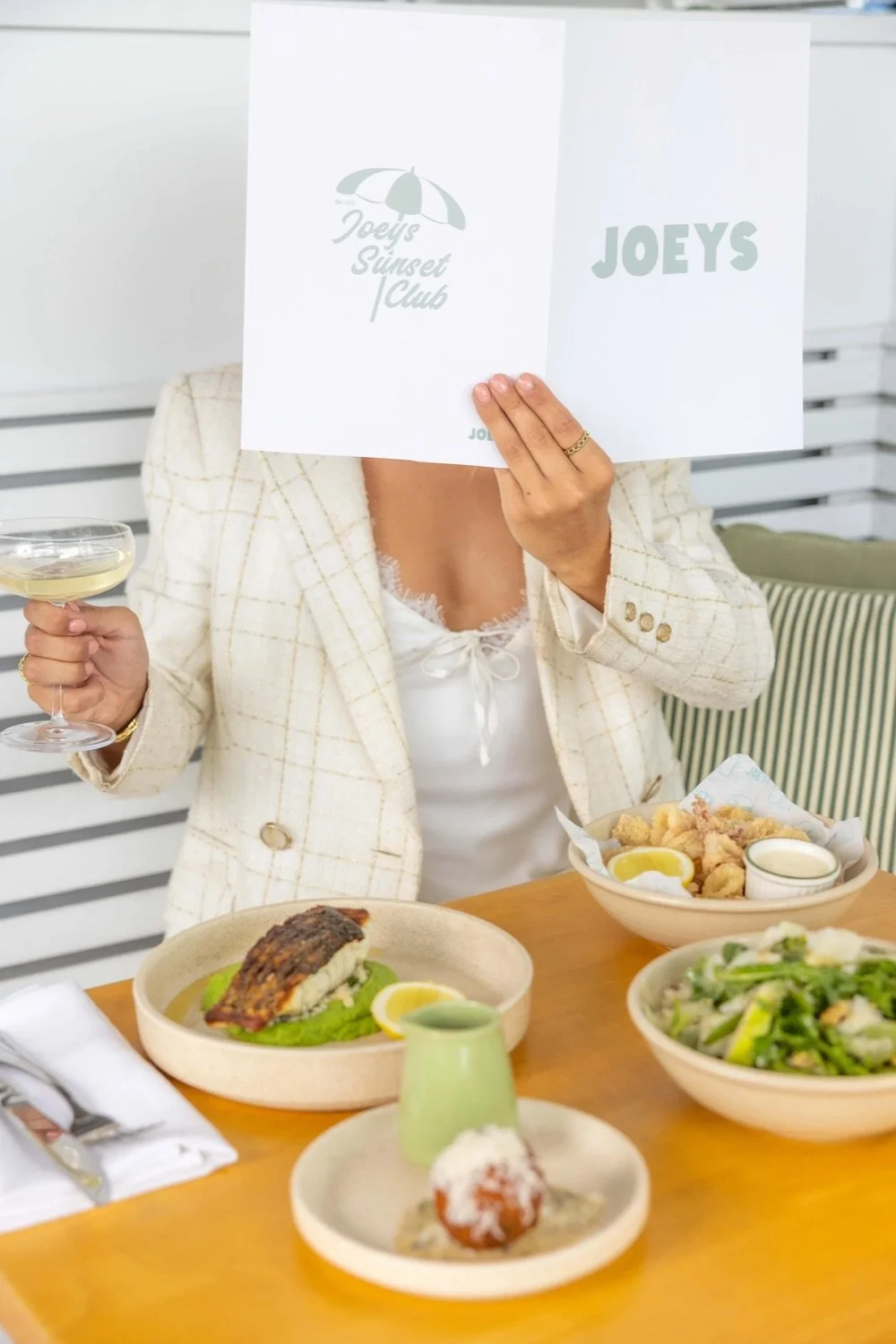 Person holding a menu in front of their face, sitting at a table with various plates of food including a piece of fish on greens, fried food with lemon, a salad, and a dessert, in a bright indoor setting.