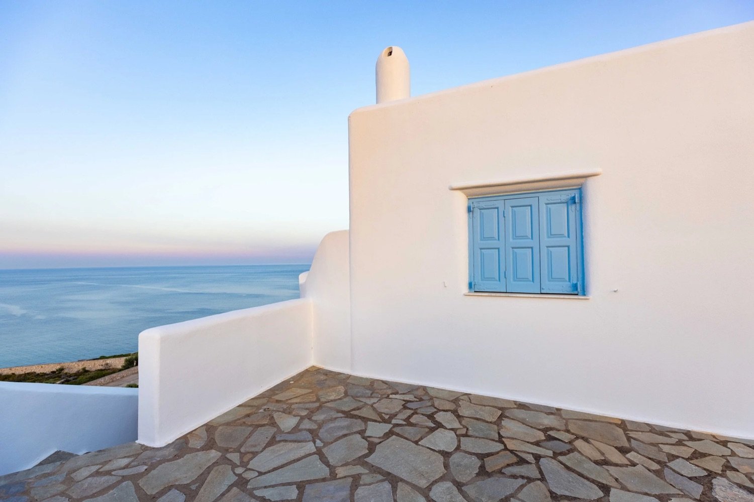 White building with a blue window shutter on a stone-paved terrace overlooking the ocean during sunset.