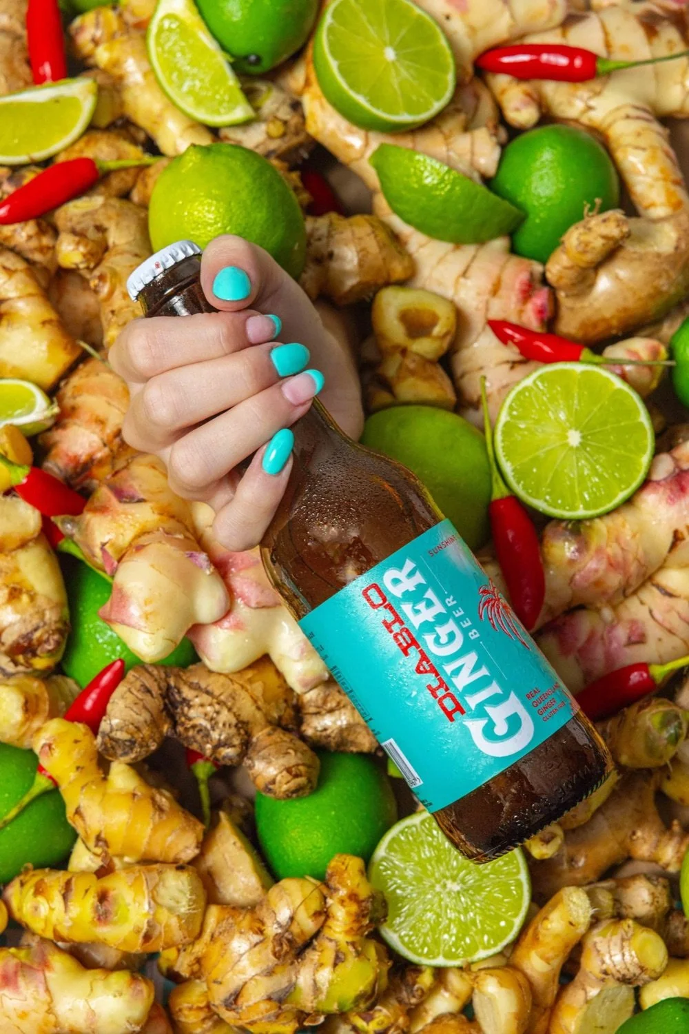 A person's hand with turquoise nail polish holding a bottle of ginger beer against a background of fresh ginger roots, green limes, and red chili peppers.