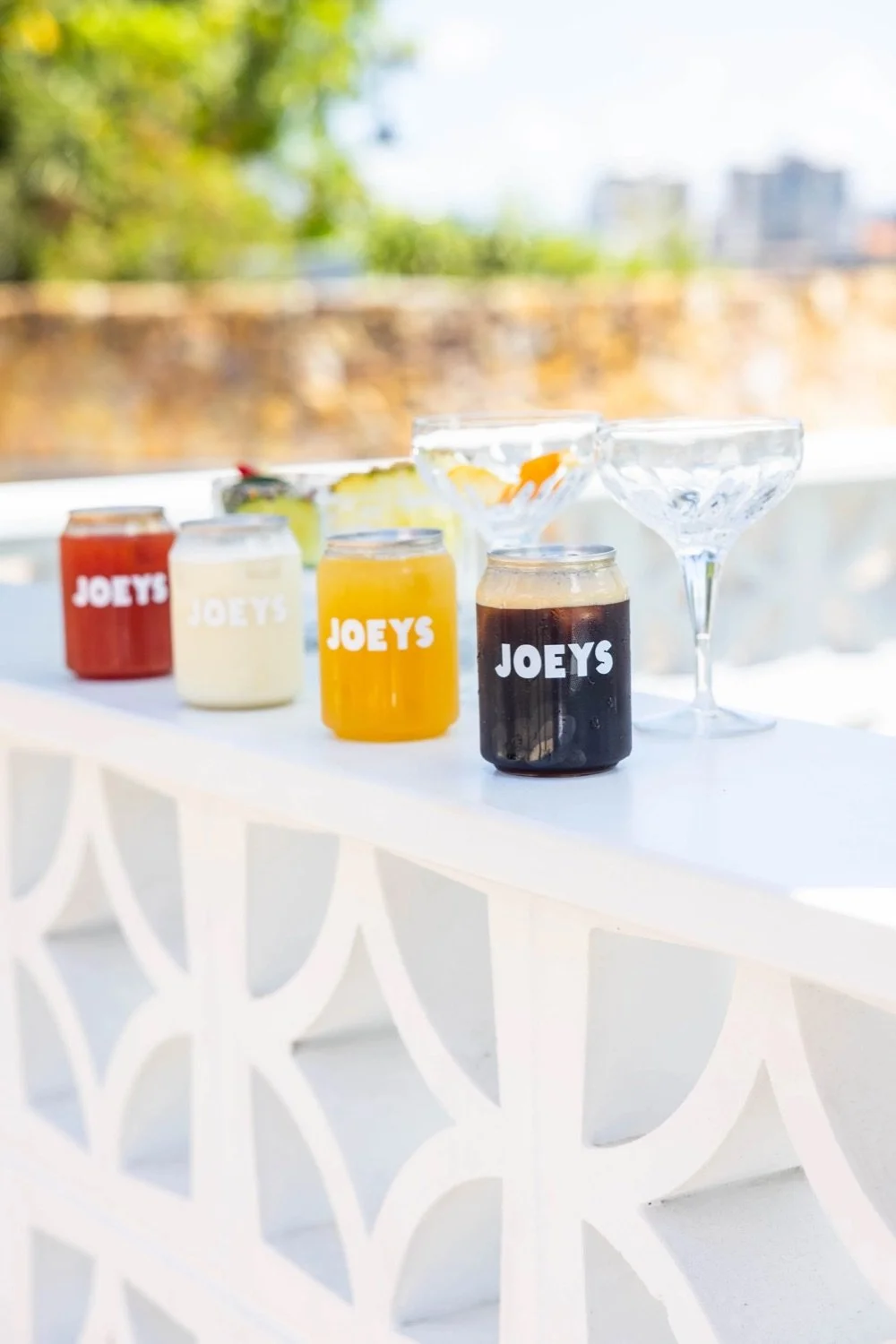 Colorful beverages in jars with 'Joey's' logo, two empty cocktail glasses, and a fruit bowl on a white outdoor table with a blurred cityscape background.