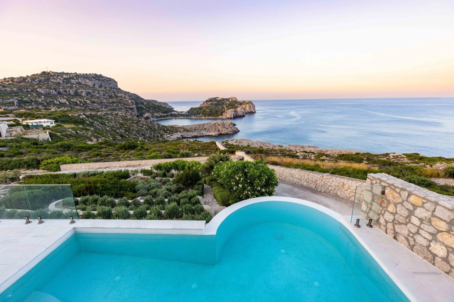 Ocean view from a villa with a turquoise swimming pool in the foreground, green bushes, rocky cliffs, and the sea at sunset.