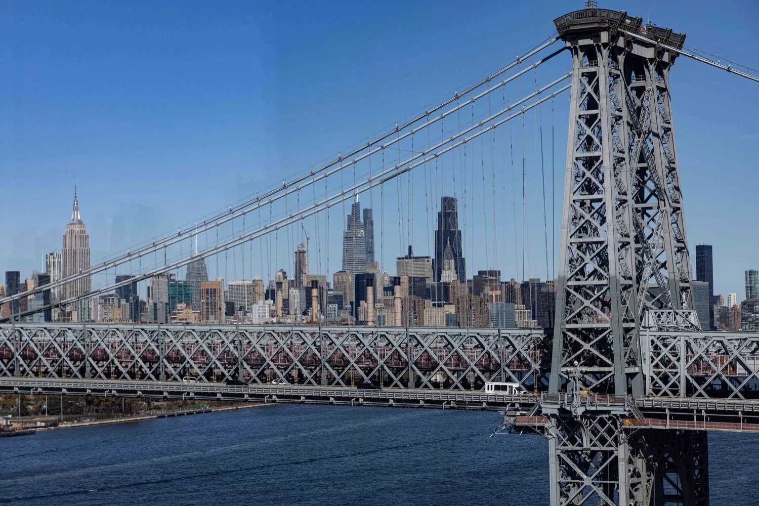 A suspension bridge with steel framework crossing a river, with the Manhattan skyline in the background, including the Empire State Building and other tall buildings under a clear blue sky.