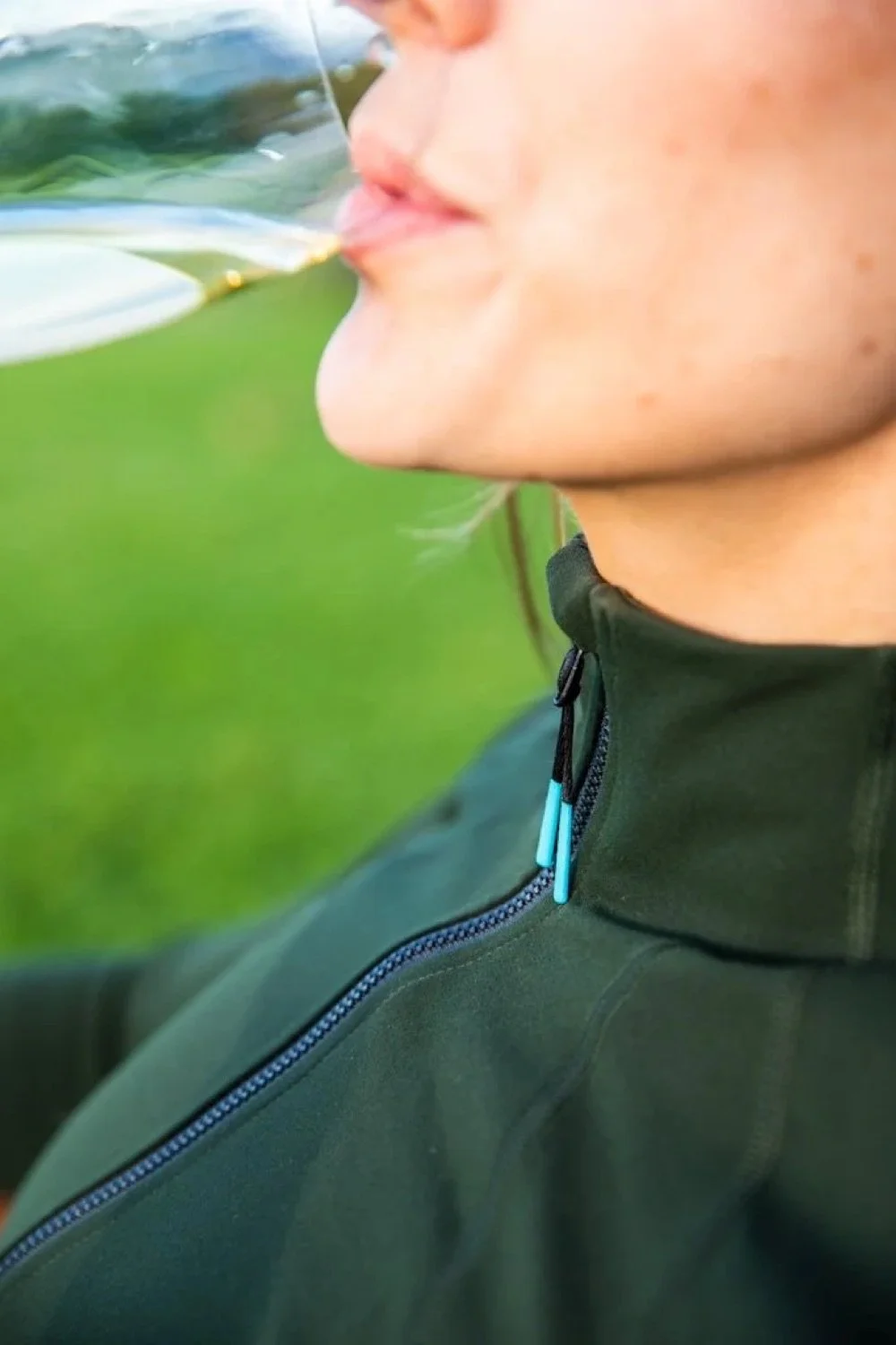 Person drinking from a clear glass with a greenish liquid outdoors, wearing a dark green jacket with a zipper.