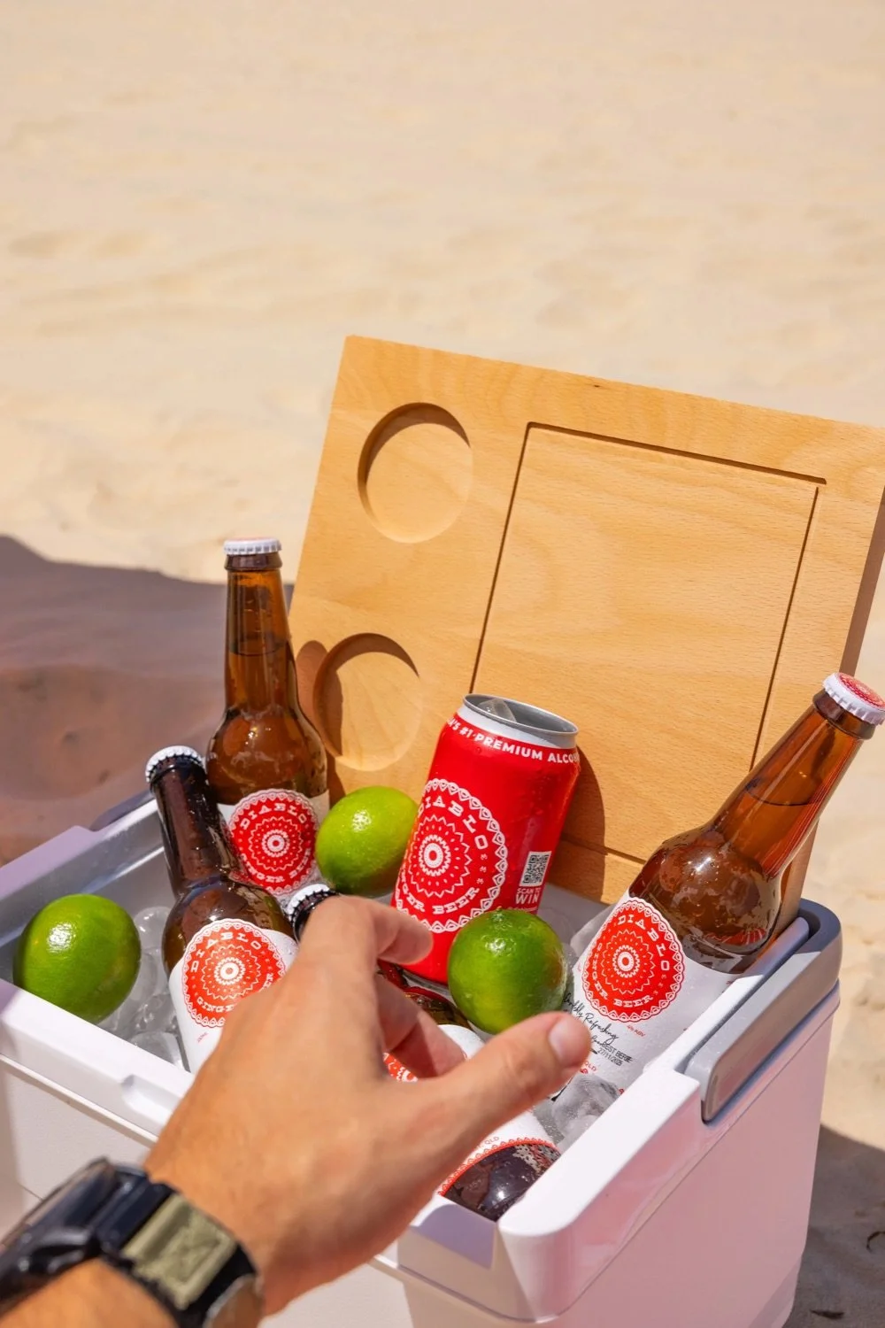A hand reaching for a lime in a cooler filled with ice, soda bottles, and beer bottles, with a wooden cutting board in the background, on a beach.