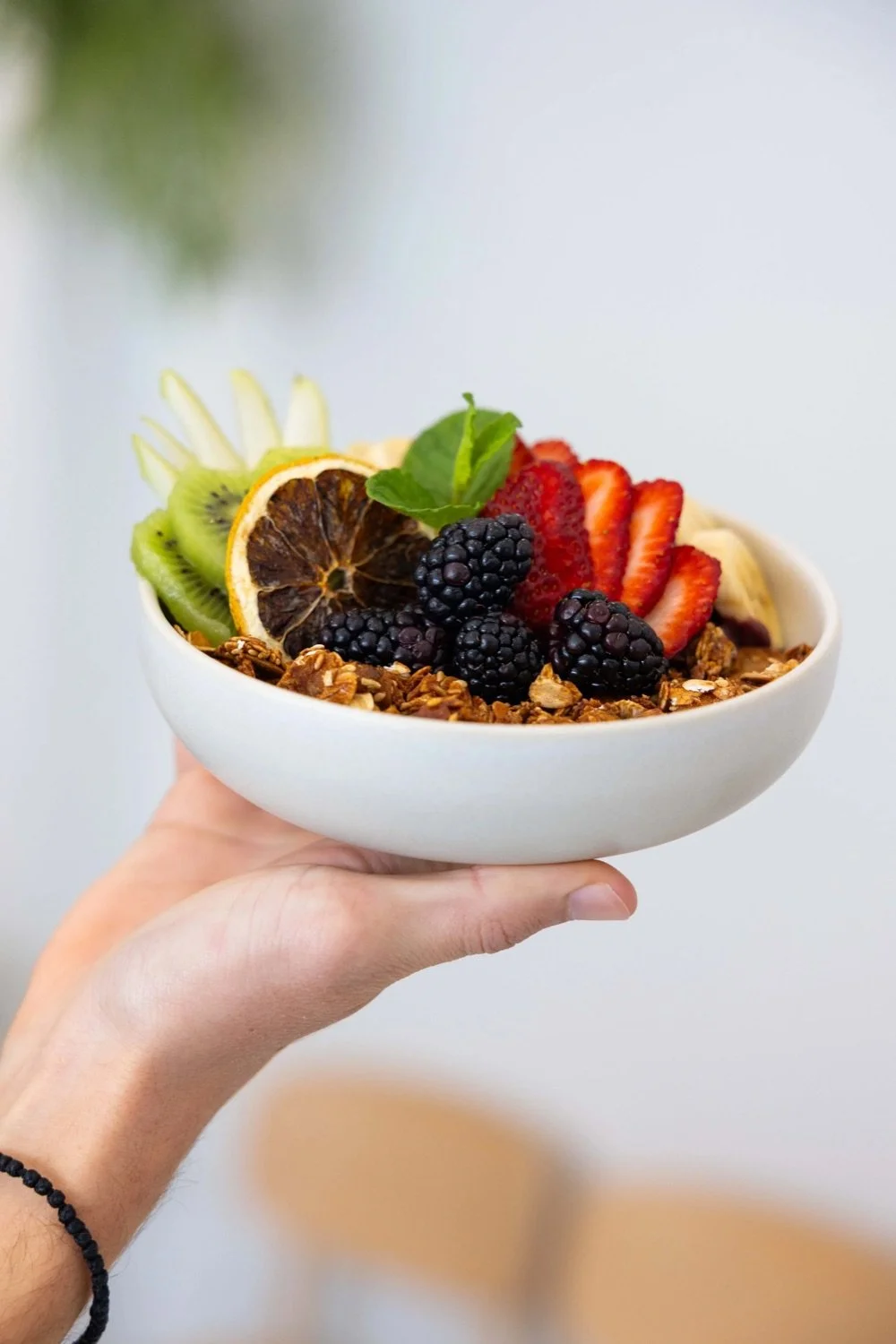 A person holding a white bowl filled with granola, topped with sliced strawberries, blackberries, kiwi, a slice of dried lemon, and a mint leaf.
