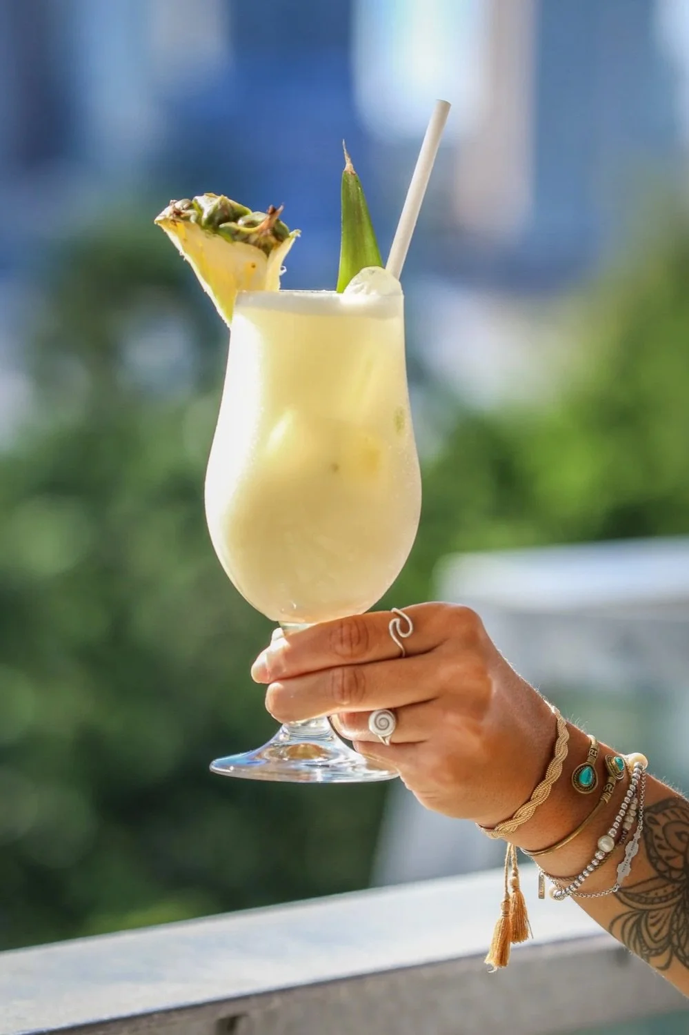 A hand holding a tall glass of pineapple cocktail garnished with a pineapple slice and a pineapple leaf.