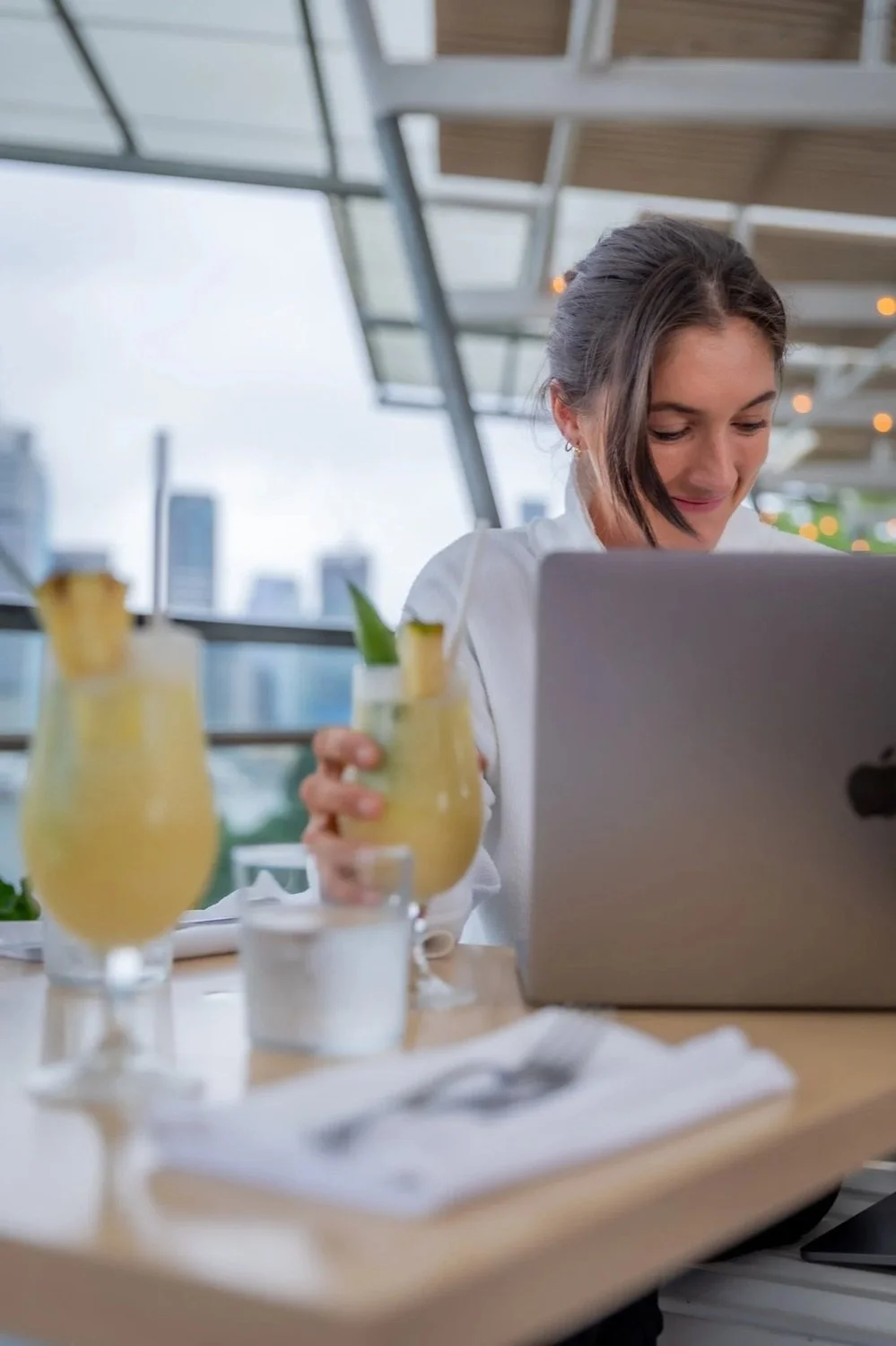 A woman in a white shirt sitting at a restaurant table, working on a laptop, with three yellow drinks garnished with pineapple and mint in the foreground, and a city skyline visible through large windows behind her.