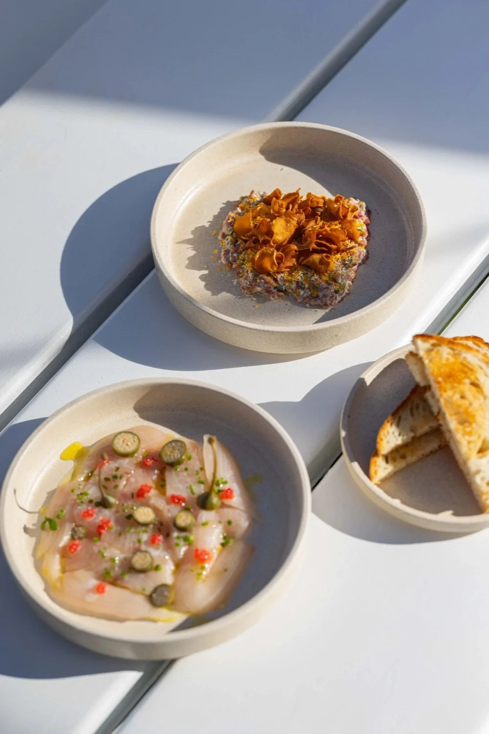 Three small bowls on a white table, one contains a slice of bread with tops optional, the second has a raw fish or seafood fillet garnished with capers and herbs, and the third has a pile of food topped with crispy fried shallots.