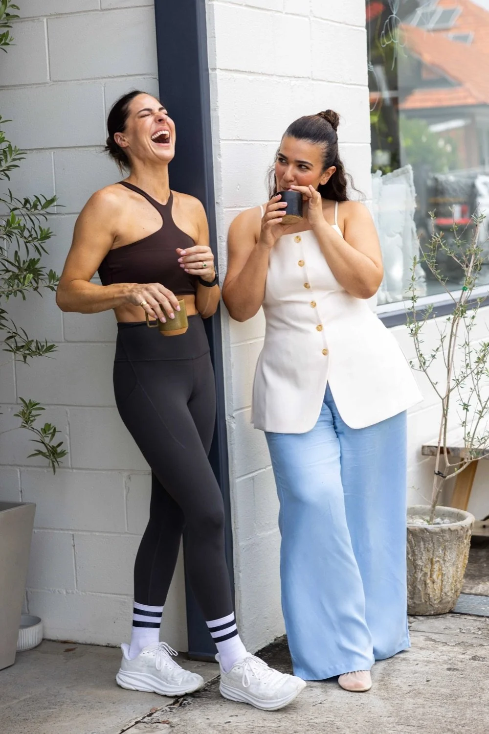 Two women are standing outside, leaning against a white brick wall, smiling and laughing. One woman is dressed in athletic wear, holding a coffee cup, the other in a white sleeveless top and light blue wide-leg pants, also holding a cup and sipping f
