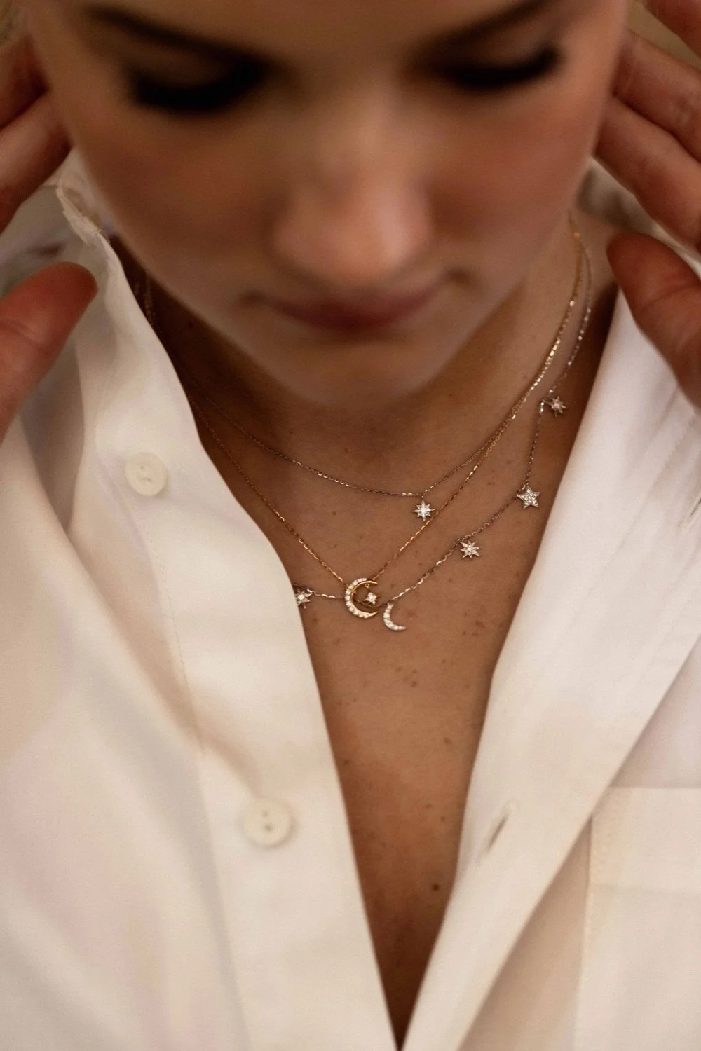 Close-up of a woman wearing layered necklaces with star and moon charms, donning a white shirt, with her hands near her ears.