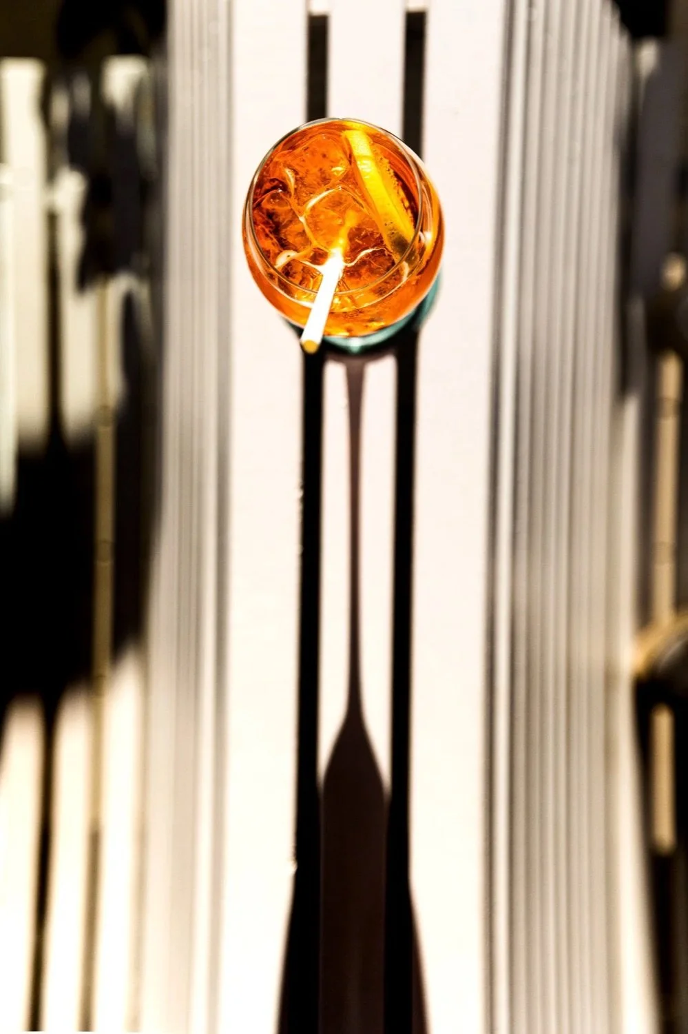 Top-down view of a drink with ice, lemon slice, and straw in a glass, casting a long shadow on a metallic surface.