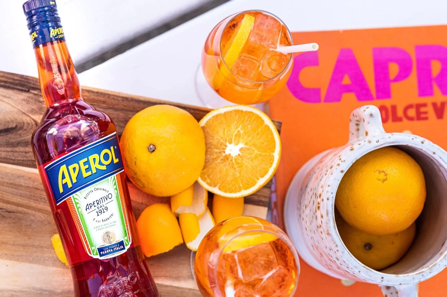 A bottle of Aperol, halved oranges, a glass of orange cocktail with ice and lemon slice, a ceramic mug with lemon and a box with vibrant pink and purple text, on a wooden surface