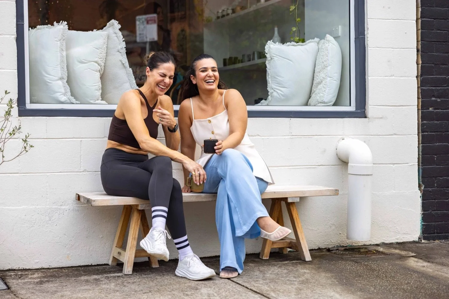 Two women sitting on a wooden bench outside a building, laughing and enjoying each other's company. One woman is wearing a black athletic outfit and striped socks, while the other is in a white top and light blue pants with heels.