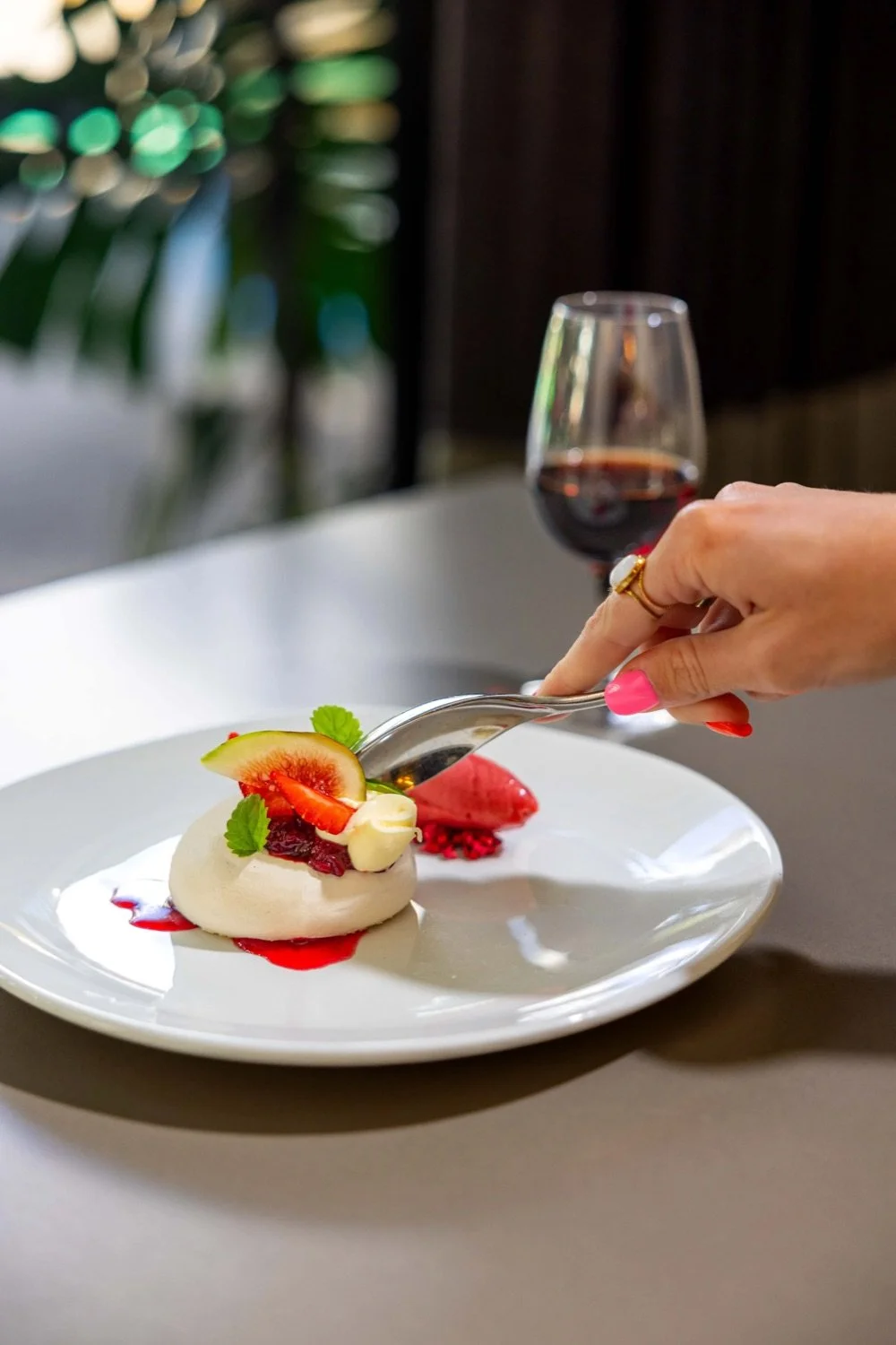 A dessert with fresh fruit, cream, and red sauce on a white plate, with a hand holding a spoon, a glass of red wine, and a background with blurred lights.