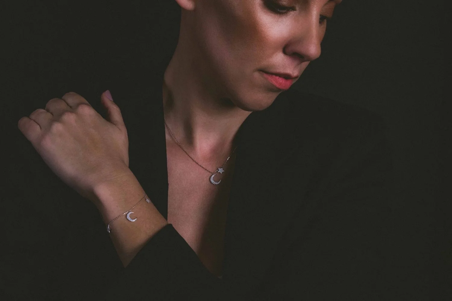 A woman with short dark hair wearing a black outfit and jewelry with crescent moon and star designs, looking downward against a dark background.