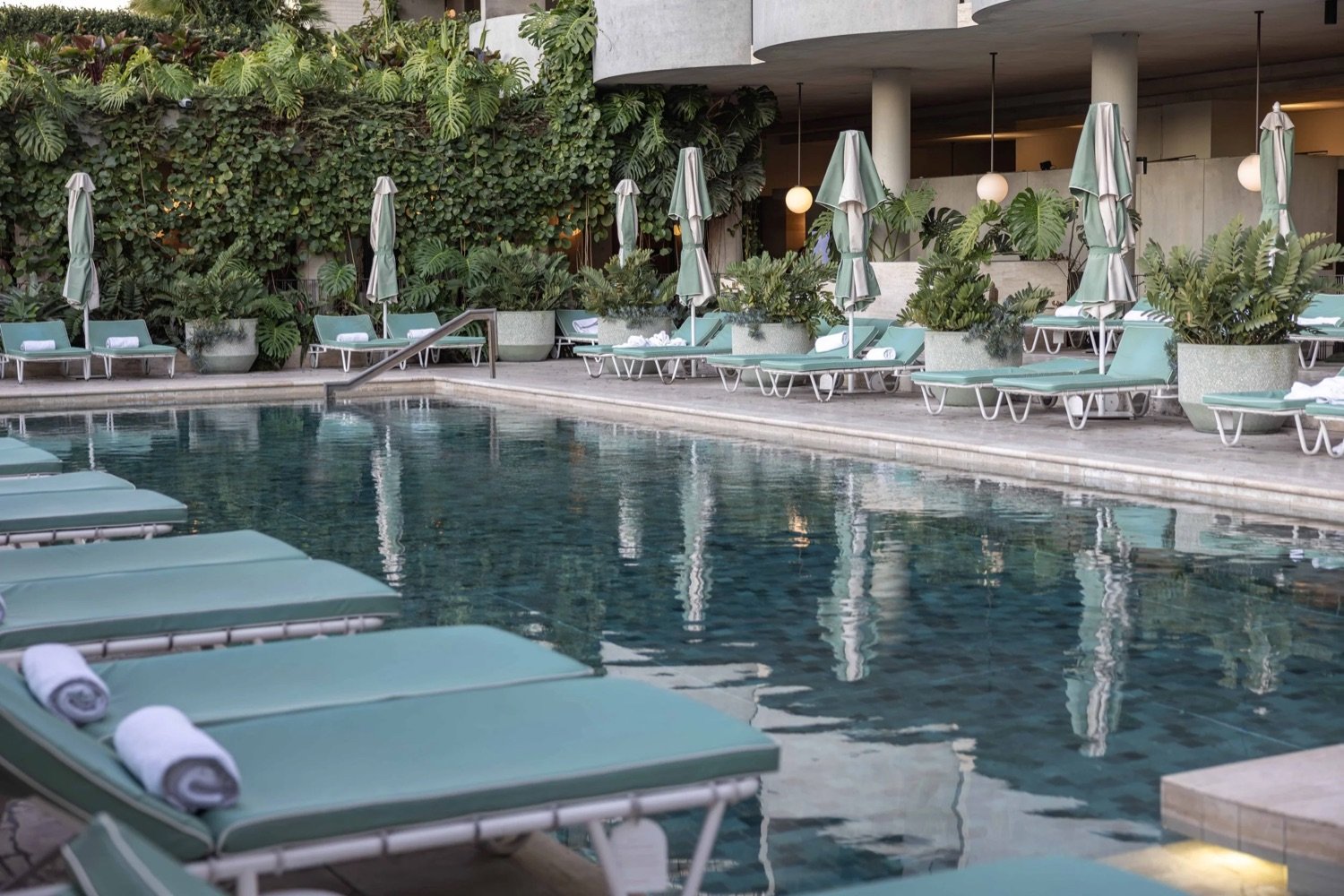 Empty swimming pool area with lounge chairs and umbrellas, lush greenery in the background, and a modern building with round lights.