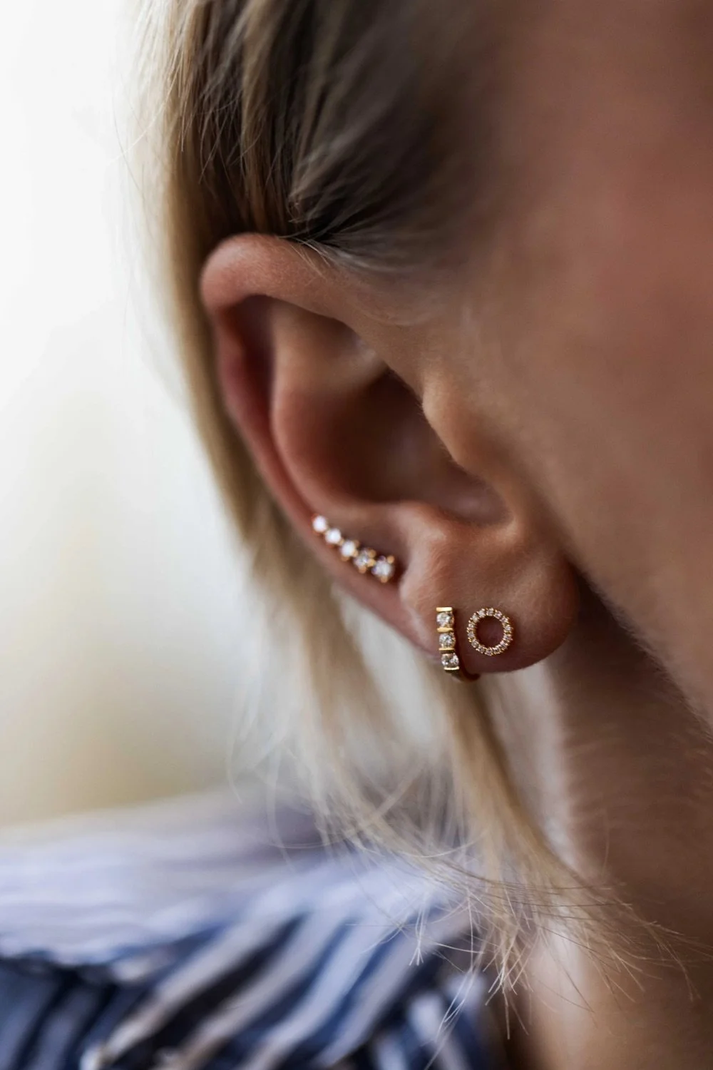 Close-up of a person's ear with multiple earrings including a small hoop with diamonds and a larger circular diamond-studded earring, with hair partially covering the ear.