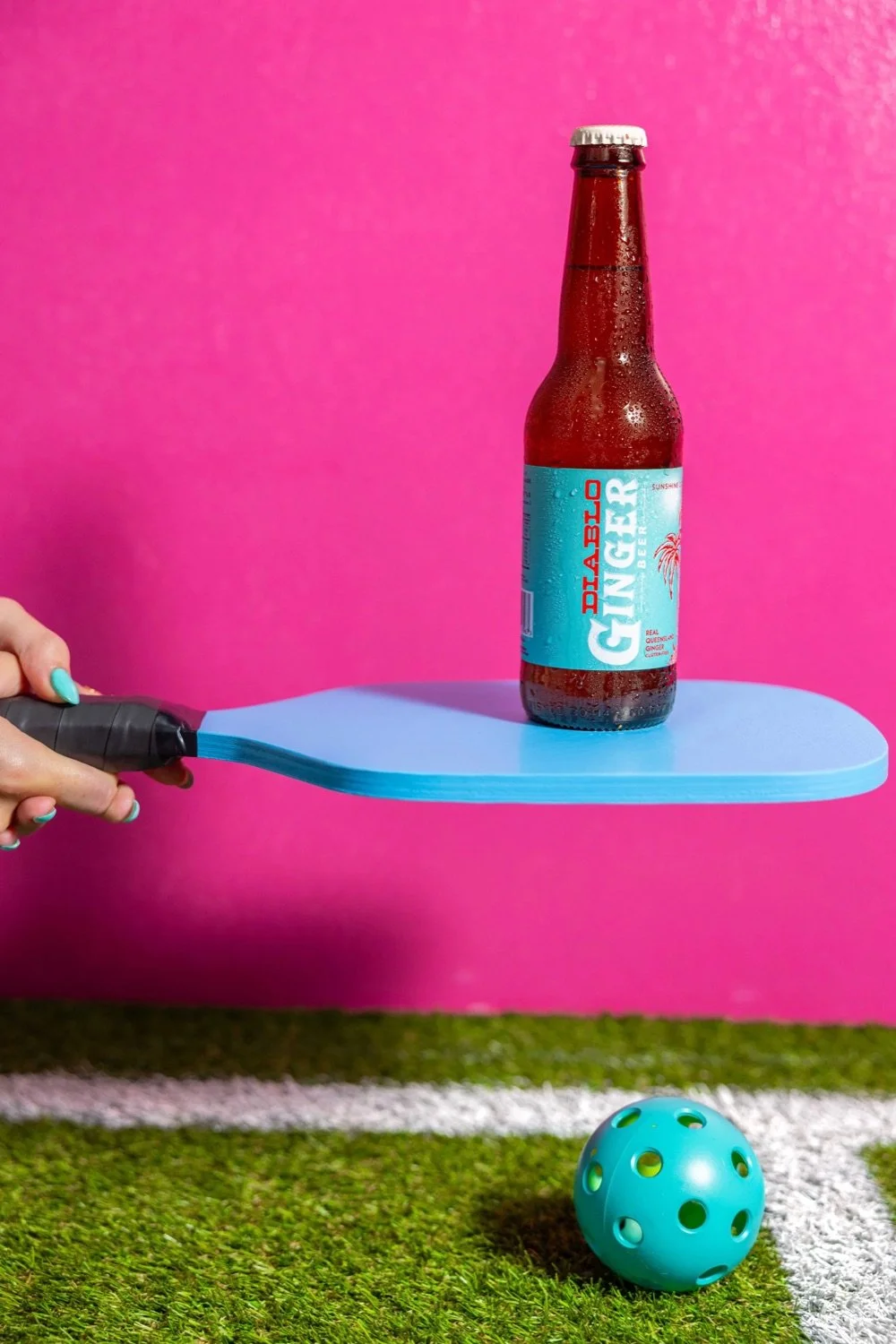 A hand holding a blue tennis racket is balancing a plastic tray with a bottle of ginger beer on it. The bottle has condensation and is labeled 'Diabolo Ginger Beer'. The background is bright pink, and there is a small blue ball with holes on a green 