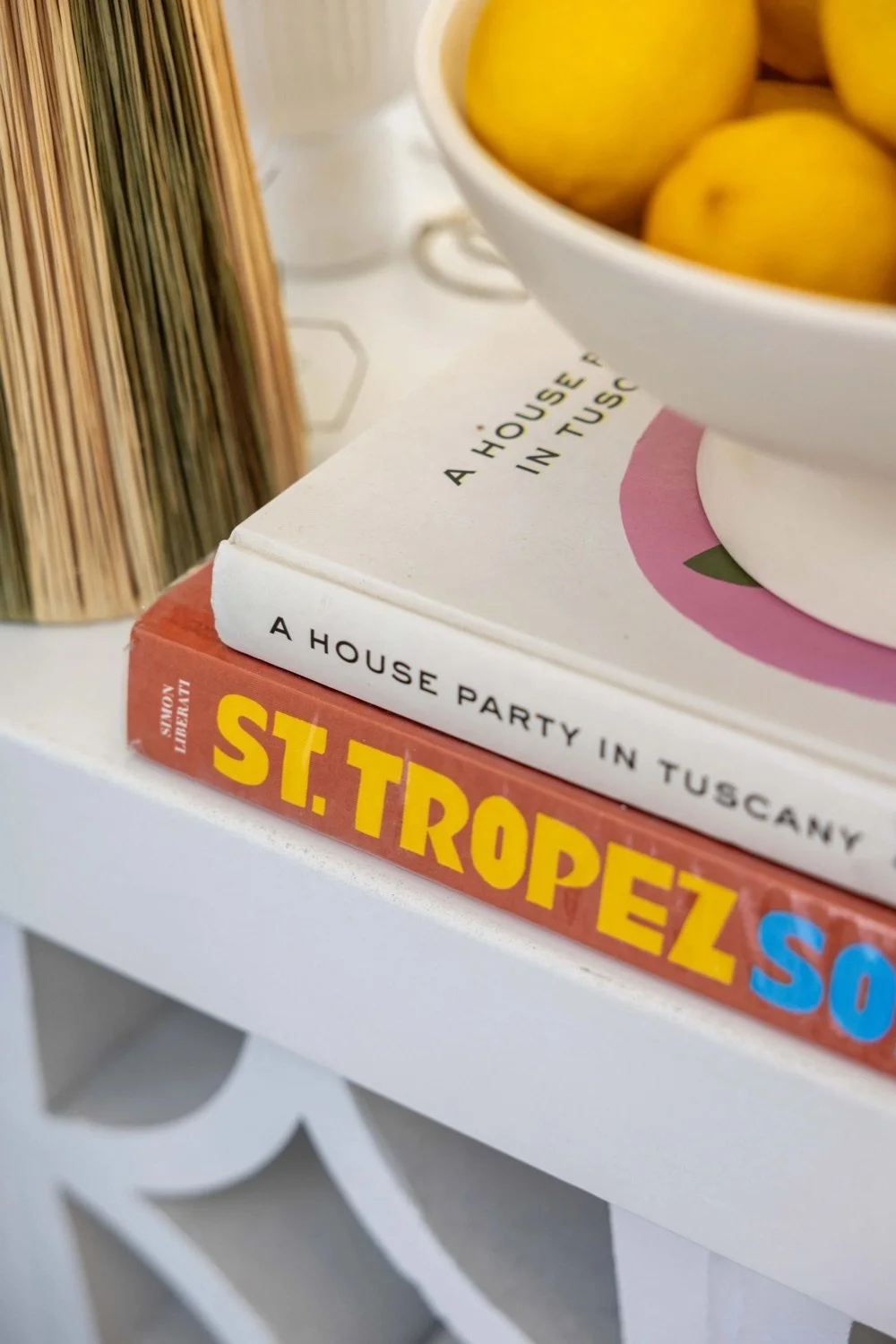 Close-up of two books stacked on a white table, with a bowl of yellow apples on top. The titles visible are "A House Party in Tuscany" and "St. Tropez". Part of a woven decorative vase is also visible.