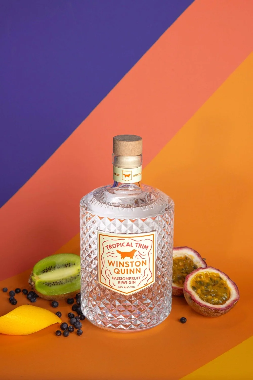 A clear textured bottle of Winston Quinn Tropical Trim Passionfruit Kiwi Gin with a wooden cap, placed on an orange surface with sliced kiwi, passionfruit, and lemon around it, against a colorful background of purple, pink, and orange.