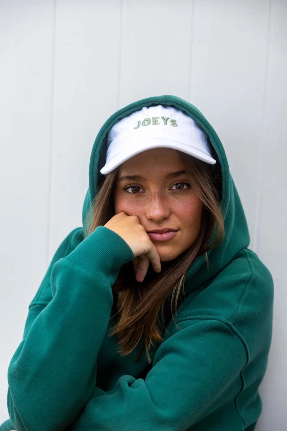 A young woman with brown hair, freckles, and light skin, wearing a white cap with 'JOEYS' embroidered on it, a teal hoodie, and sitting against a light-colored wall.