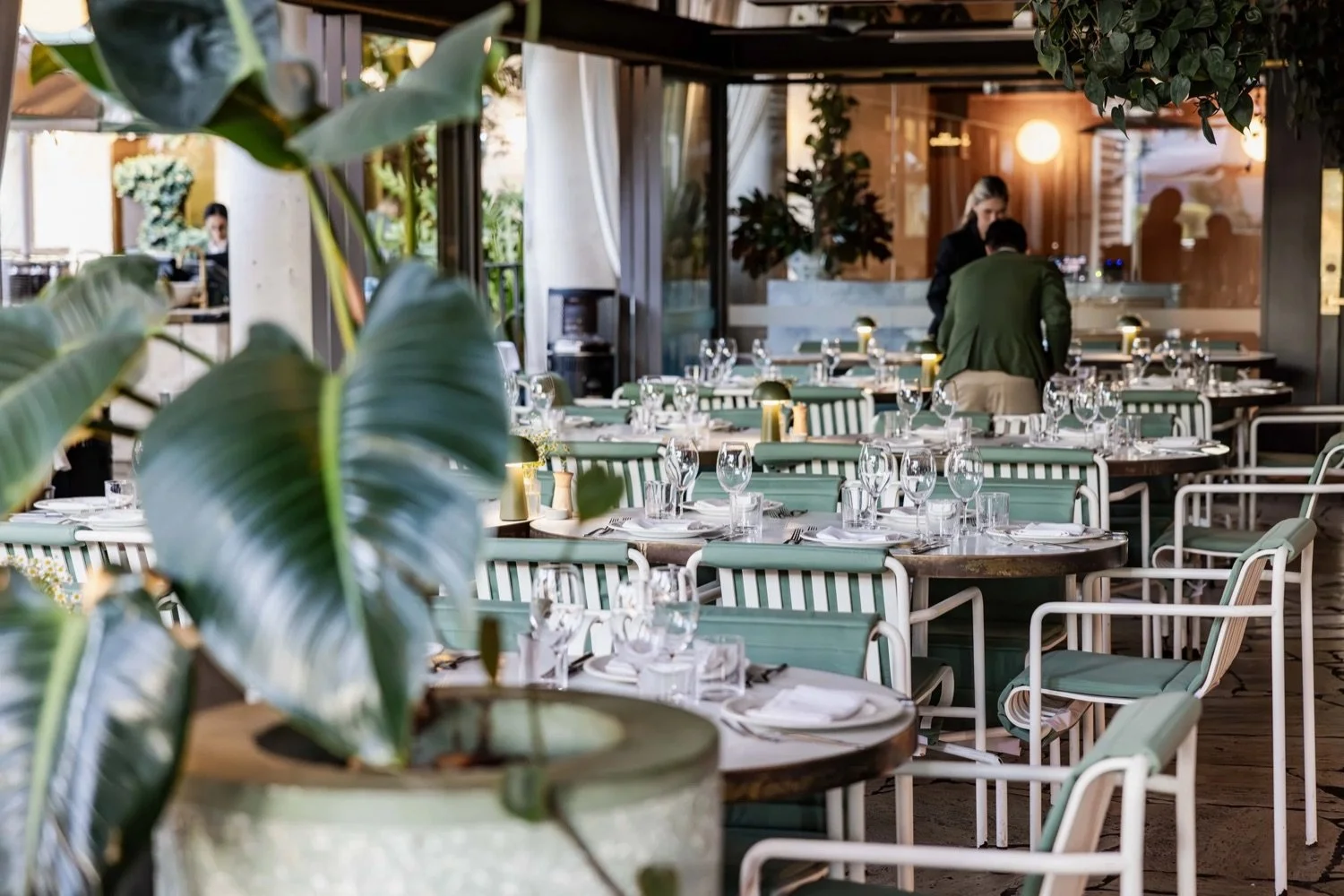 An outdoor restaurant patio with tables set with glassware, silverware, and napkins, decorated with plants, and with people preparing the area.